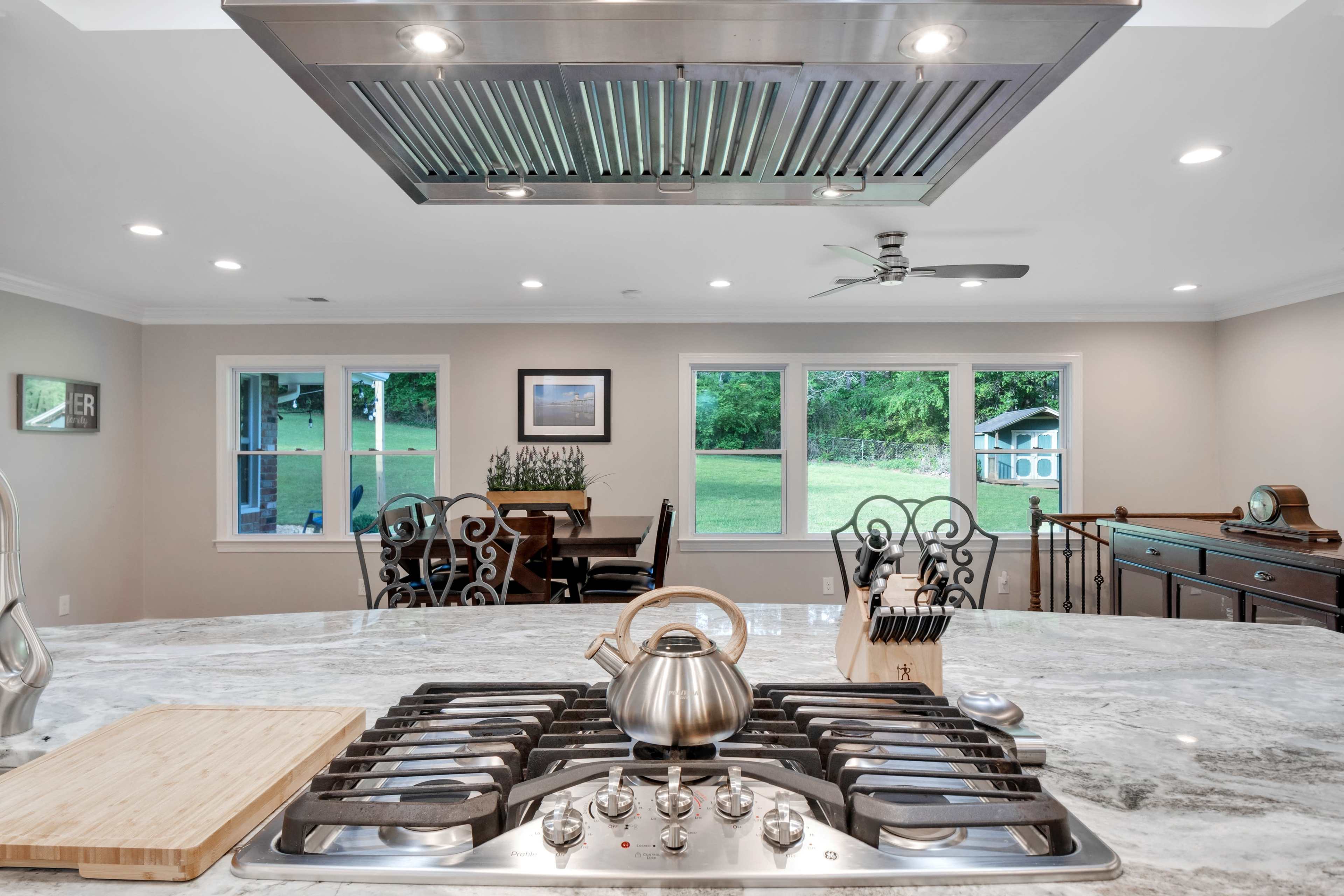 A kitchen with a granite countertop features a stainless steel stove and an overhead vent, overlooking a dining area with large windows that provide a view of the outdoors.