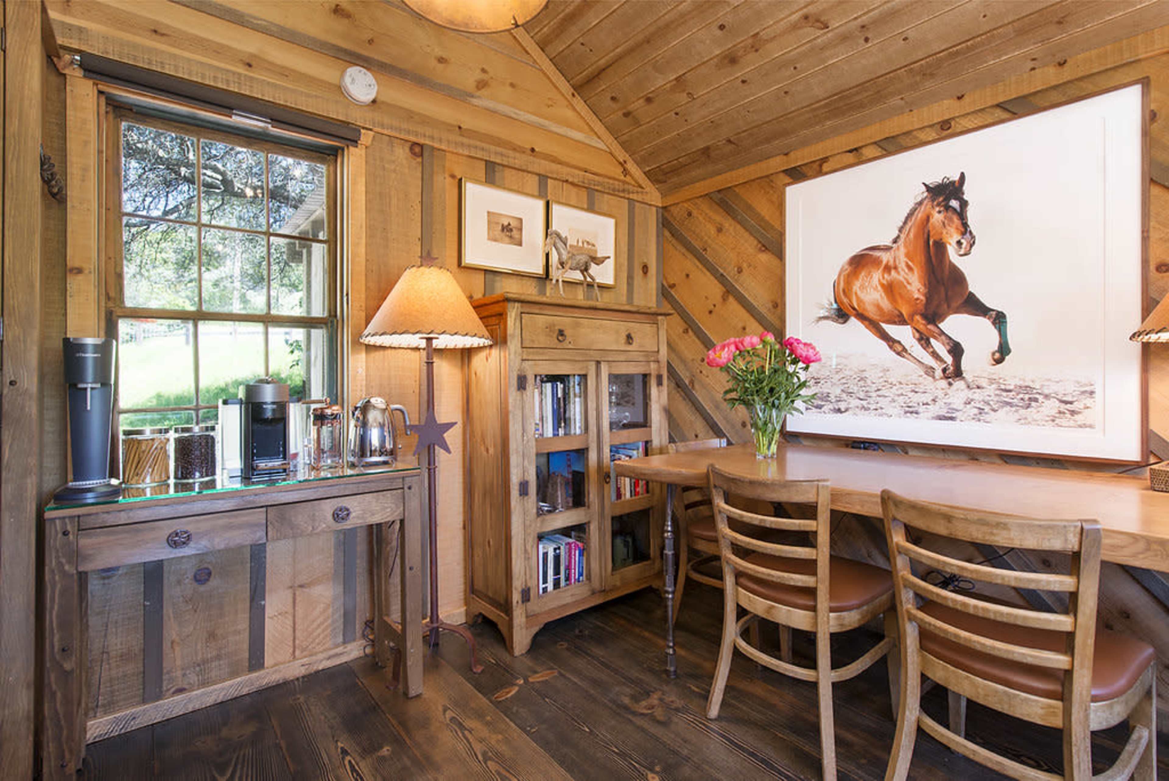 The image features a wooden interior of a small cottage with a dining table, chairs, a cabinet, and framed artwork of a horse on the wall.