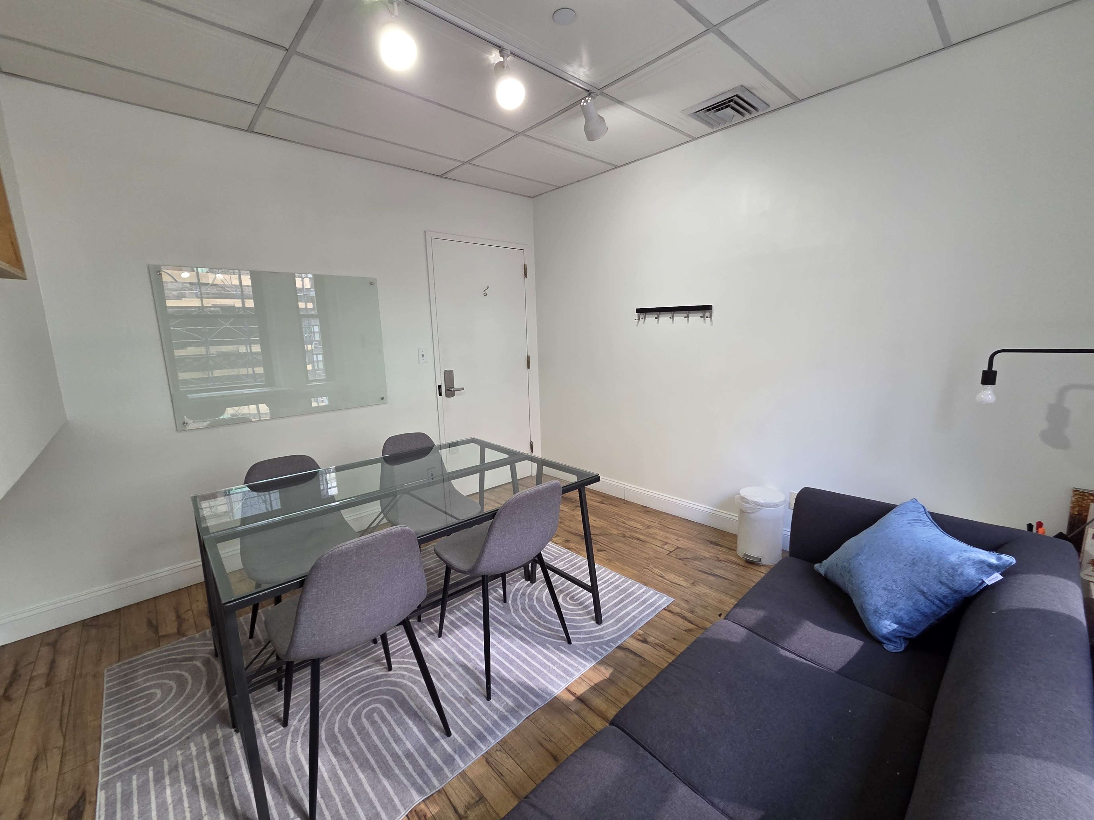 The image shows a modern, minimalist room with a dining table and four chairs, a gray sofa, and a wall-mounted coat rack.