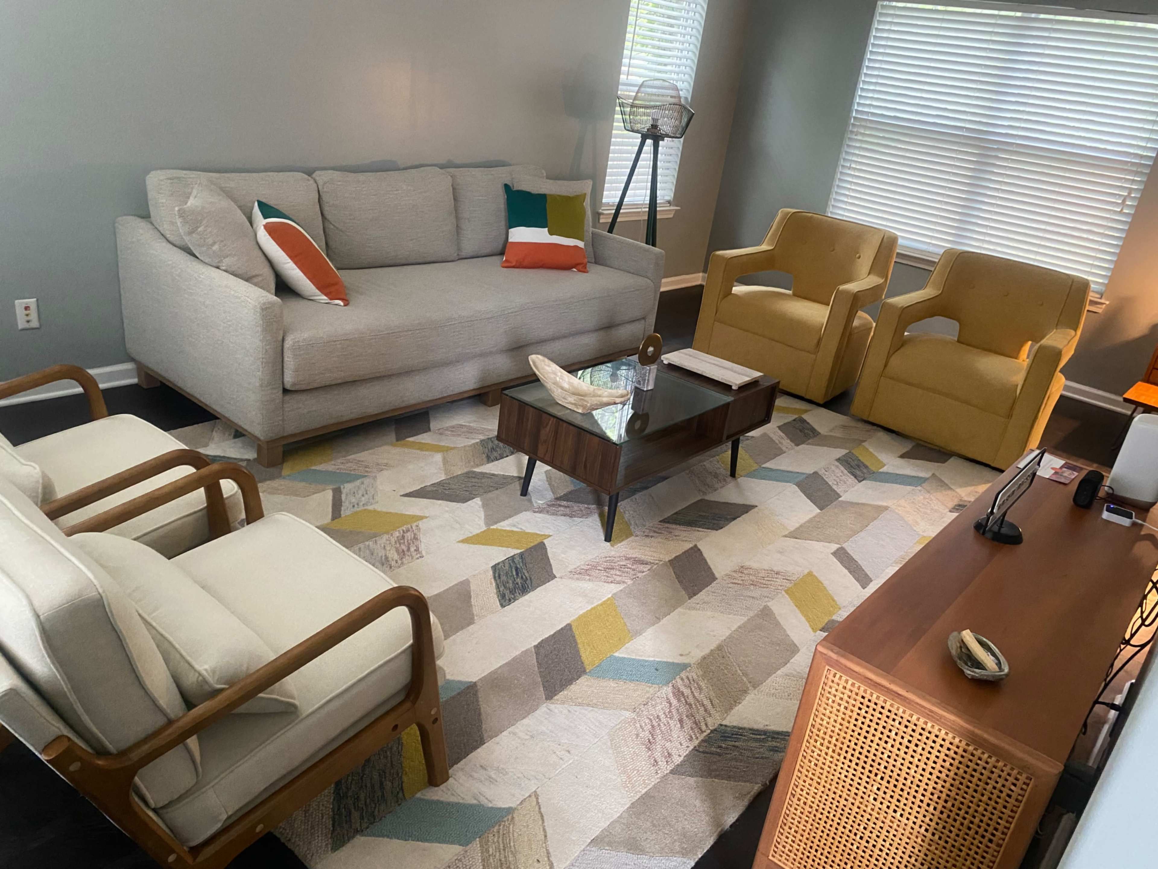 The living room features a light gray sofa, two yellow armchairs, a wooden coffee table, and a patterned area rug, with large windows letting in natural light.