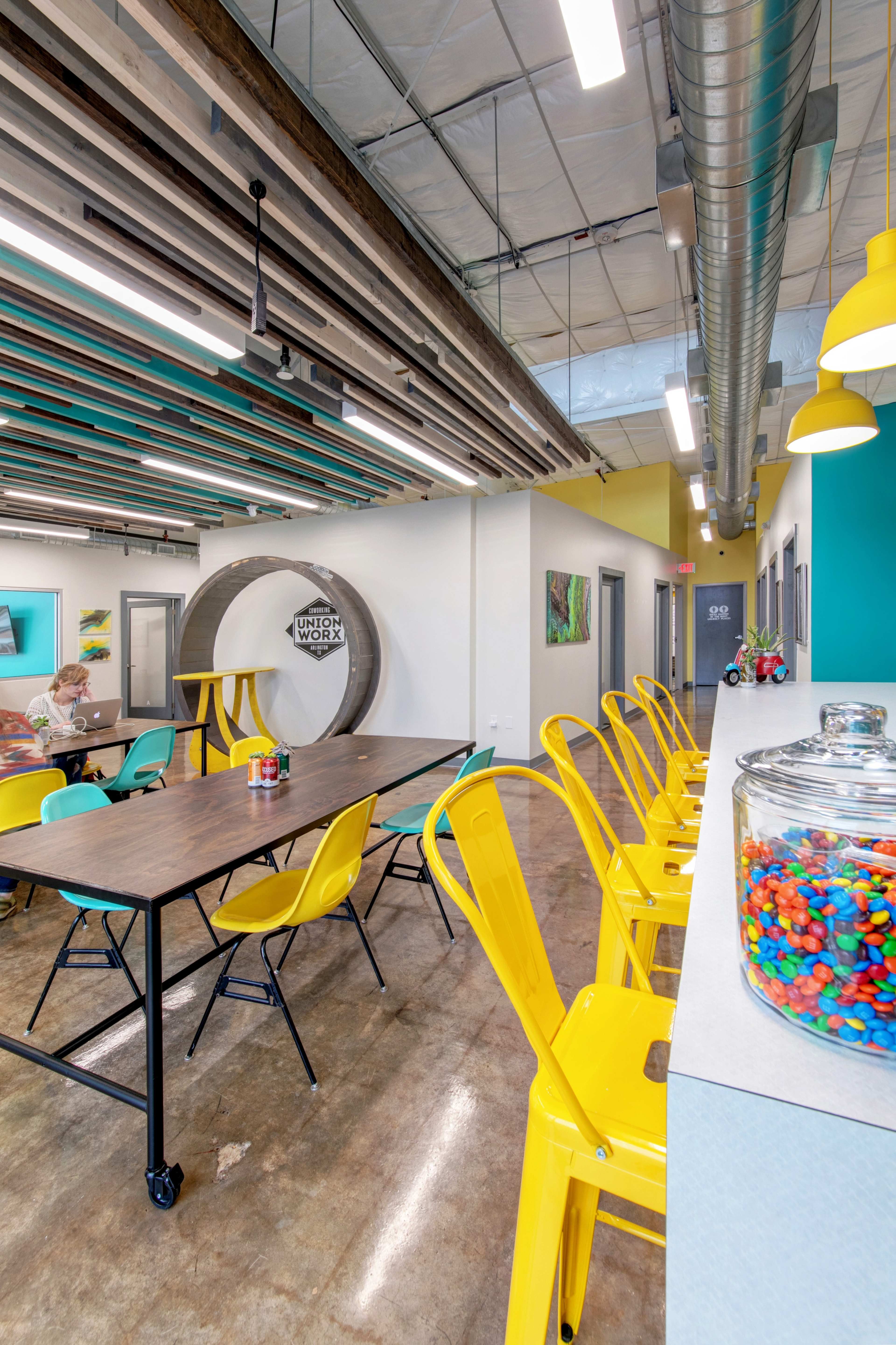 A modern workspace features a long table surrounded by yellow chairs, with a glass jar of colorful candy on a counter in the foreground.