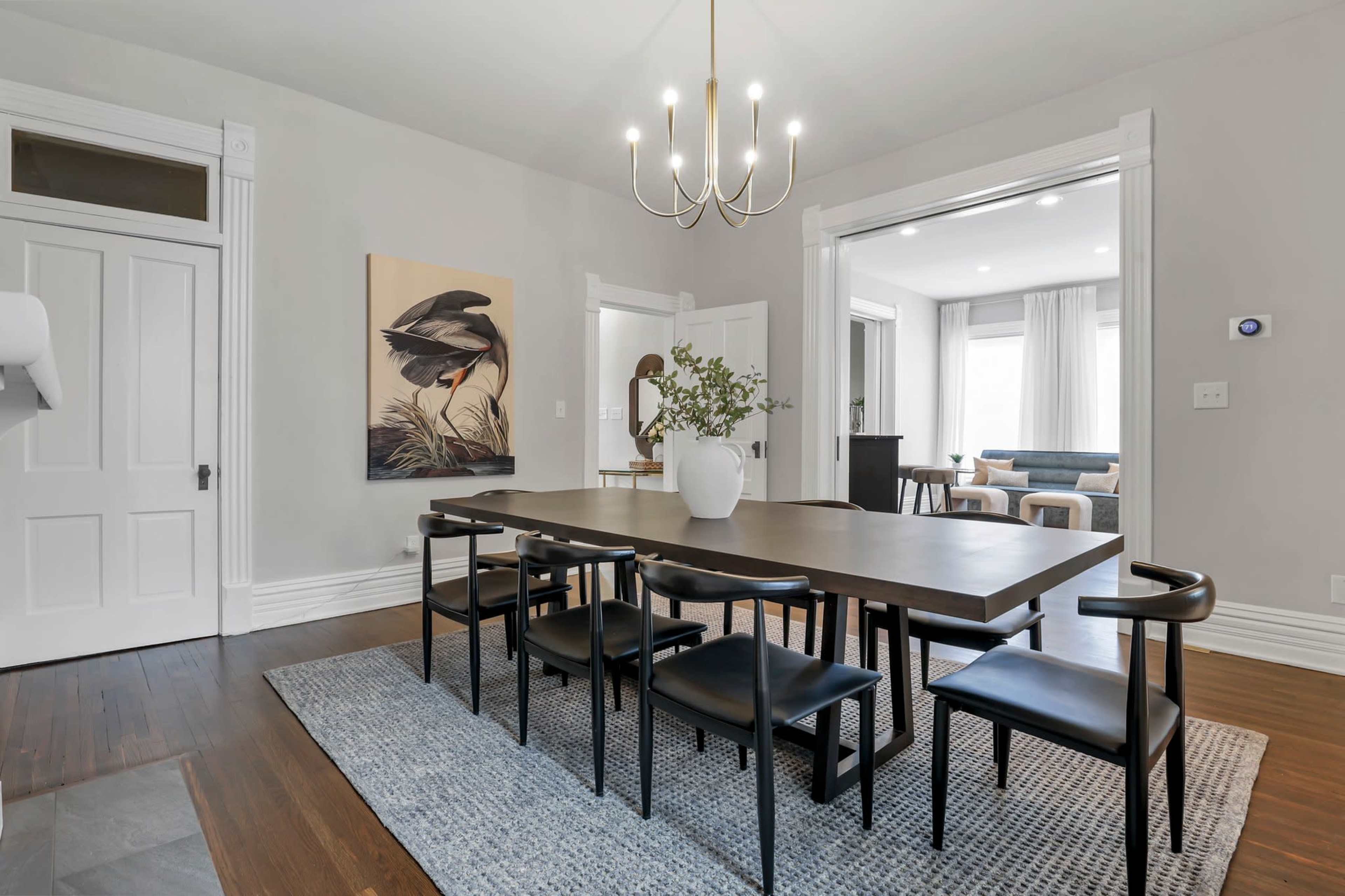 A large dining table with black chairs is positioned on a rug in a gray-walled room, featuring a wall art piece of a bird and a doorway leading to another room.