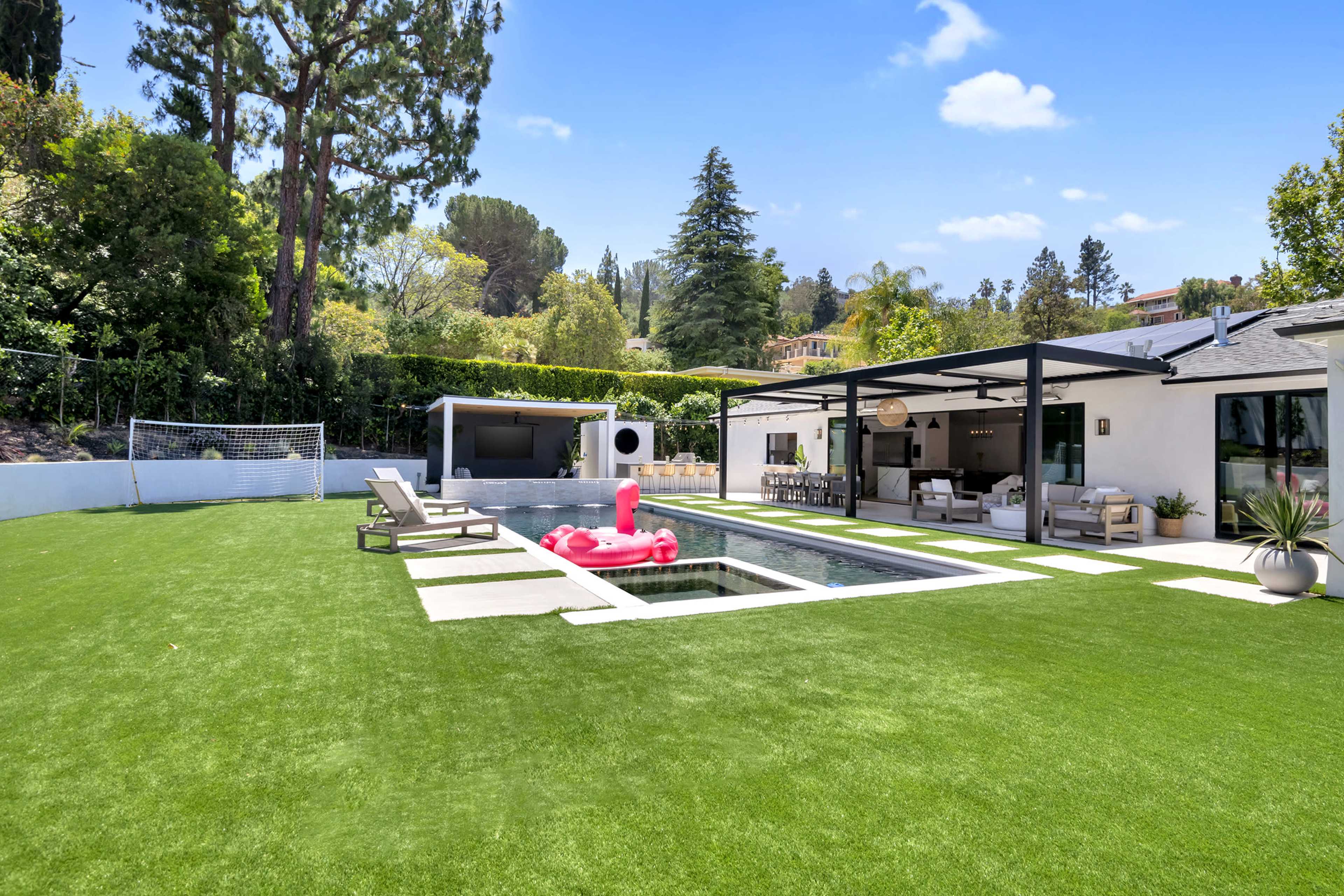 A modern backyard features a swimming pool surrounded by artificial grass, lounge chairs, and a dining area, with a goalpost in the background.