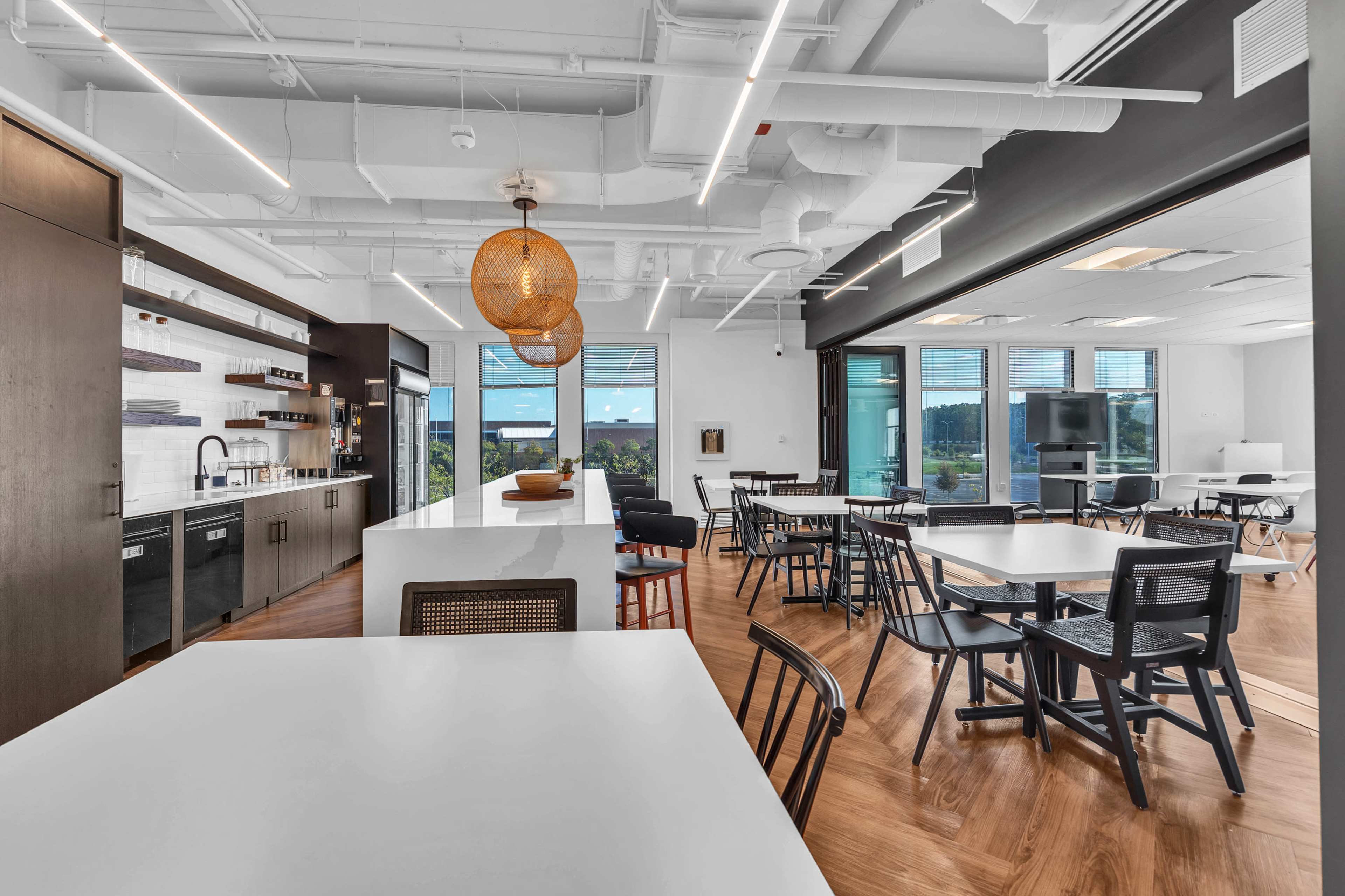 The image shows a modern office kitchen and dining area with wooden floors, a sleek white countertop, and a variety of seating arrangements.