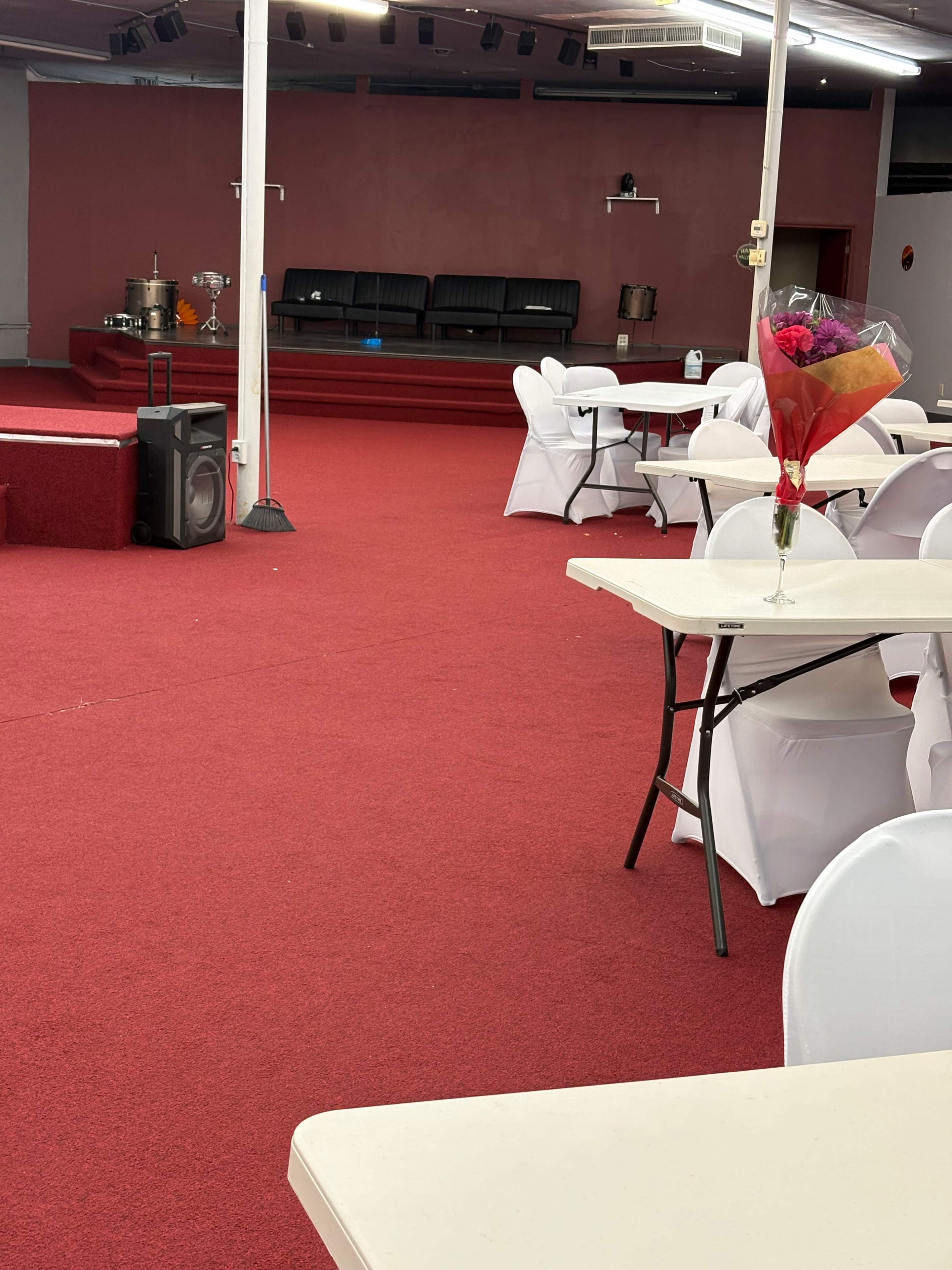 The image shows a spacious event venue with red carpet flooring, several white tables and chairs arranged around, and a raised stage at the far end.