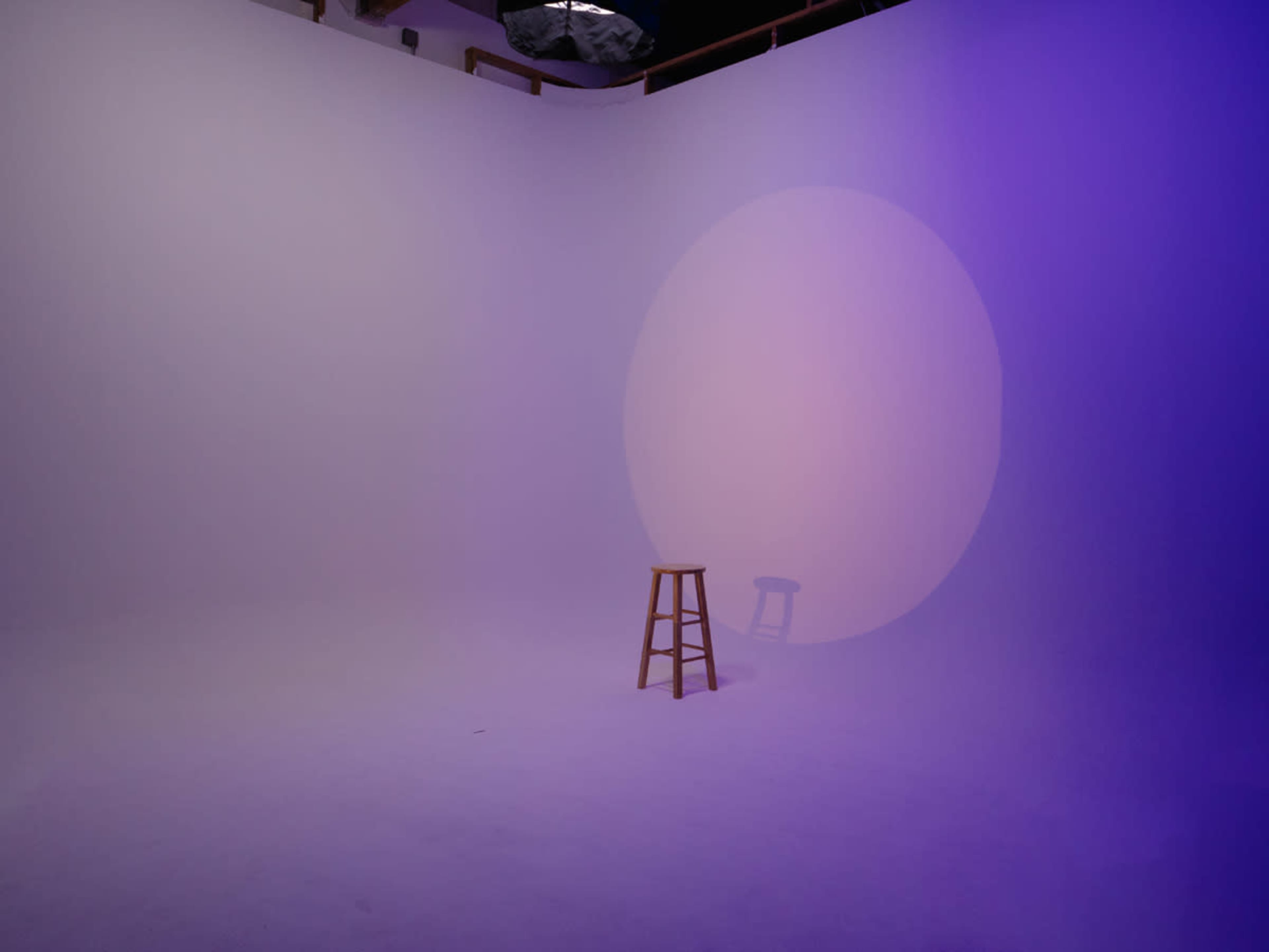 A wooden stool sits in a minimalistic photography studio with a softly lit purple backdrop.