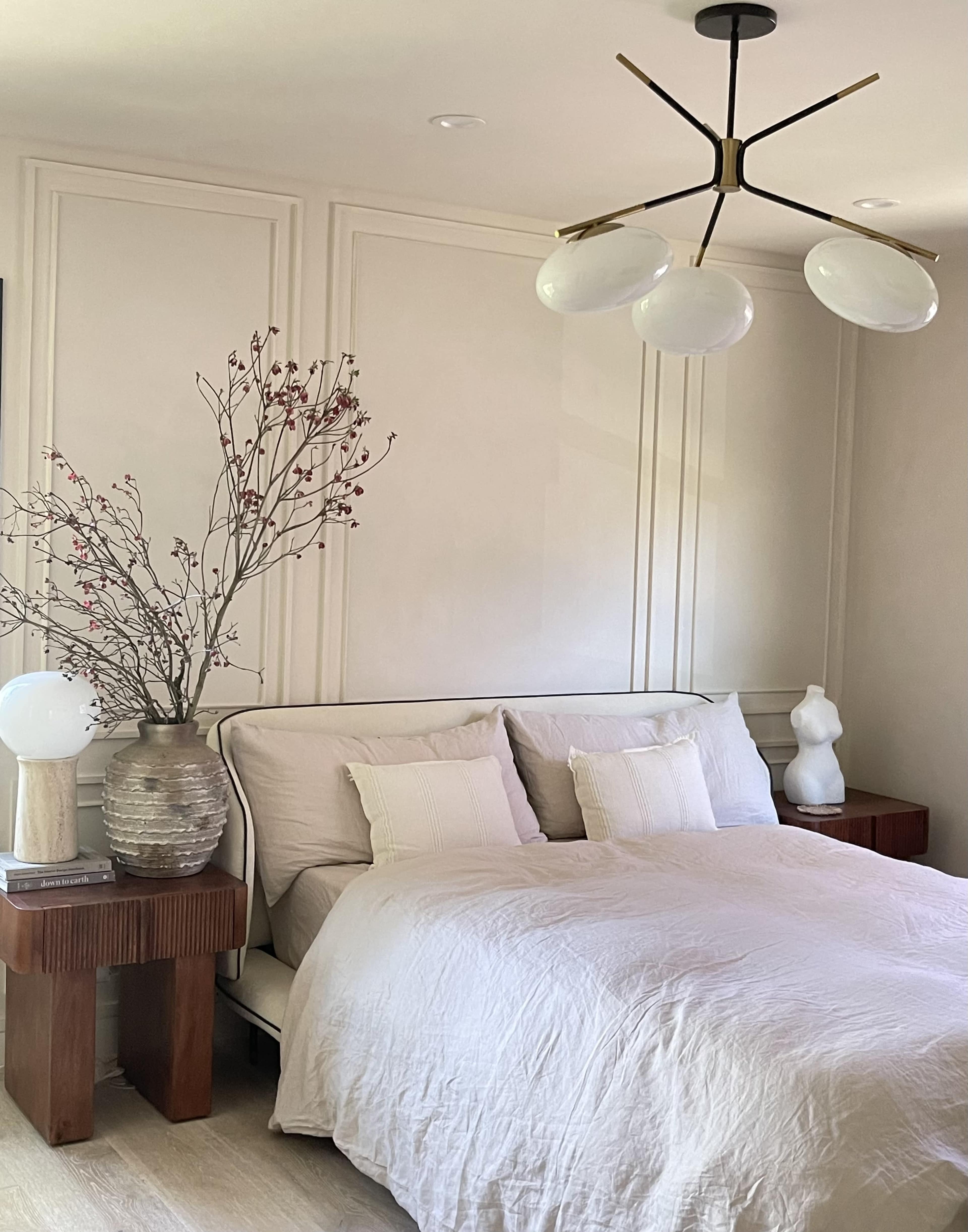 A bedroom with a bed dressed in a light-colored coverlet, flanked by wooden nightstands, a decorative vase with branches, and a modern light fixture overhead.