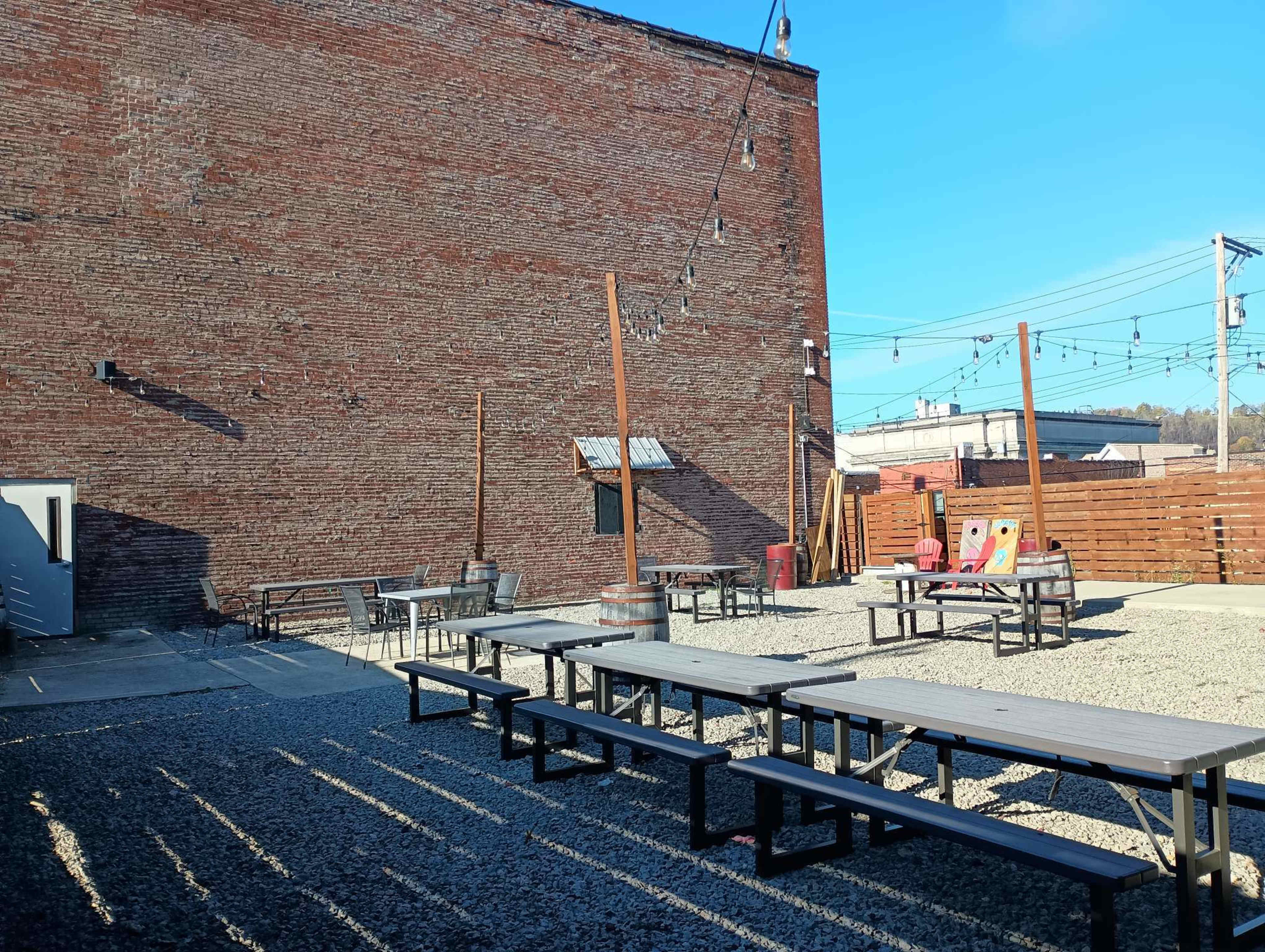 An outdoor seating area with several picnic tables and benches set on gravel, against a brick wall and under clear blue skies.