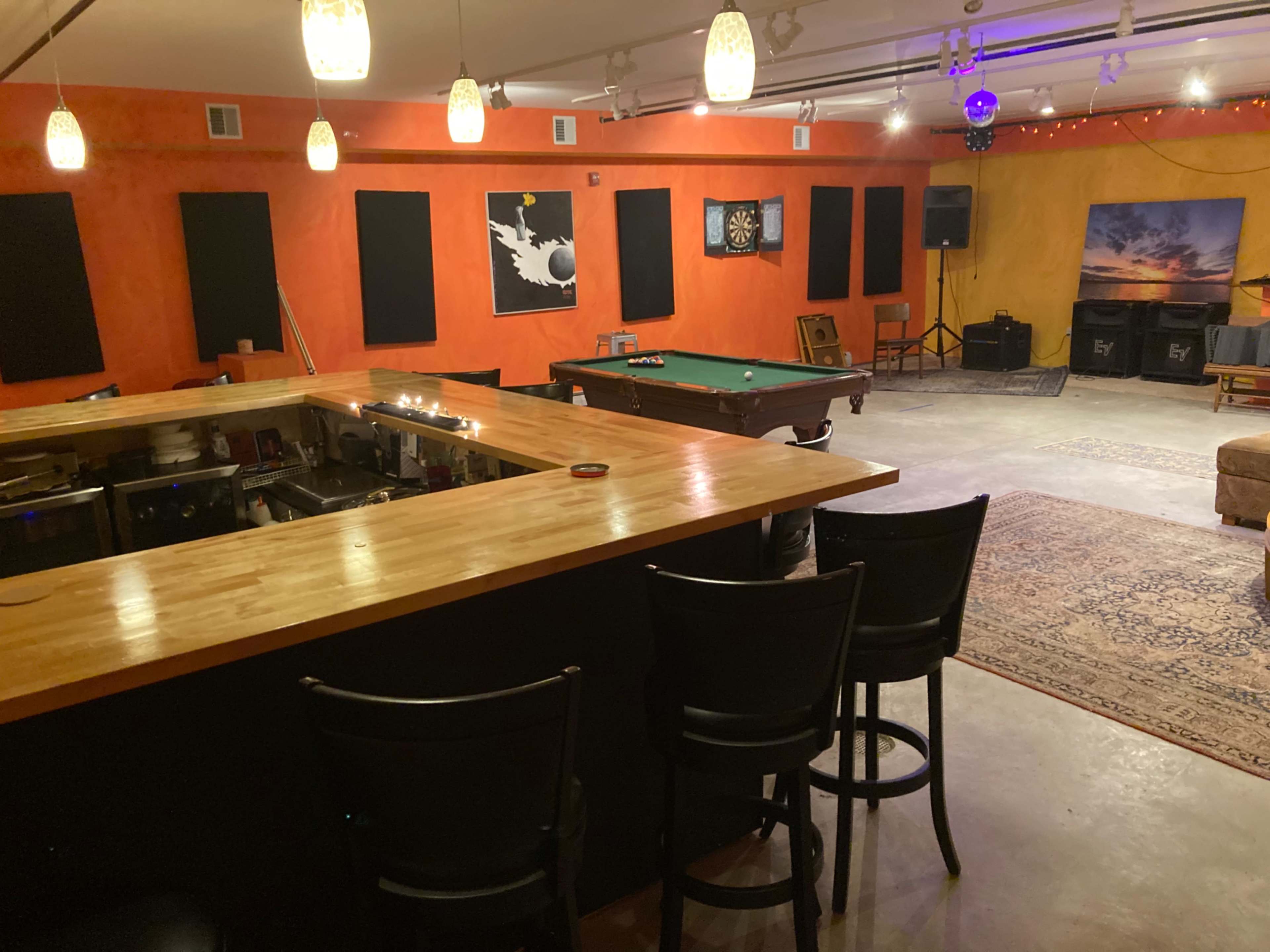 A spacious recreational room with a bar area, a pool table, and an arrangement of wall-mounted art, illuminated by overhead lights.