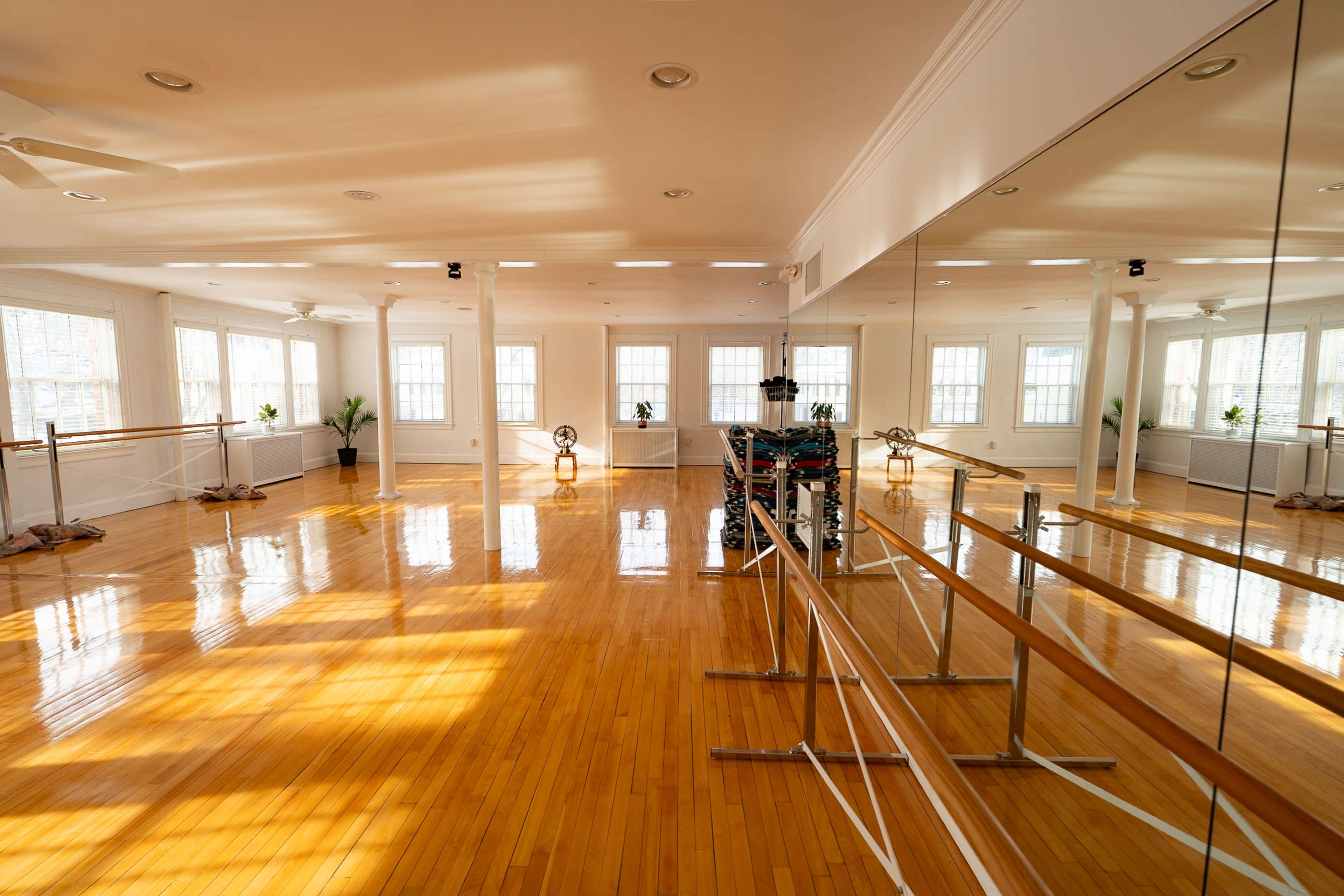 The image shows a spacious dance studio with wooden floors, large mirrors, and ample natural light streaming through the windows.