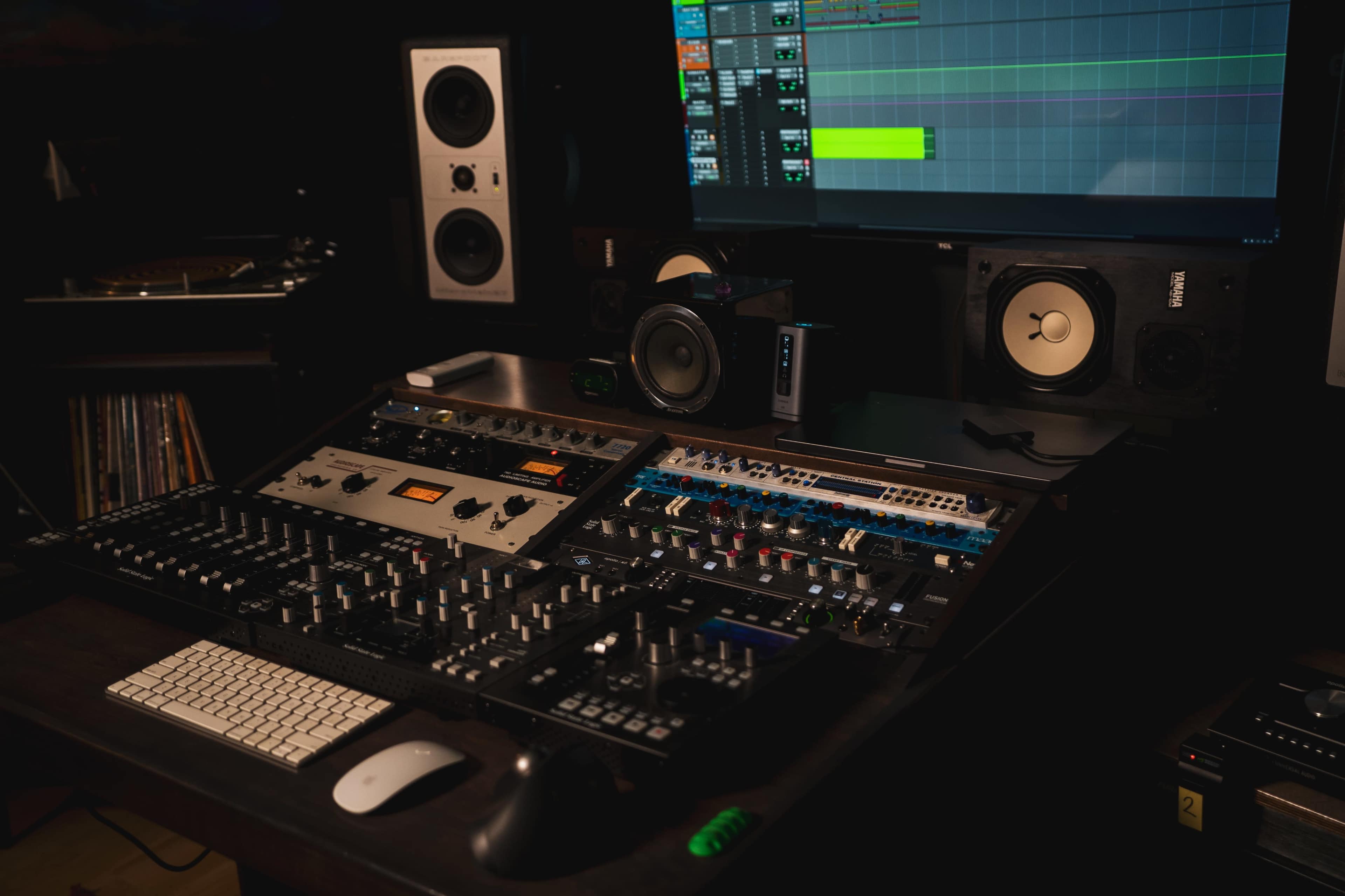 The image shows a music production studio with a large mixing console, audio equipment, and a computer monitor displaying a digital audio workstation.