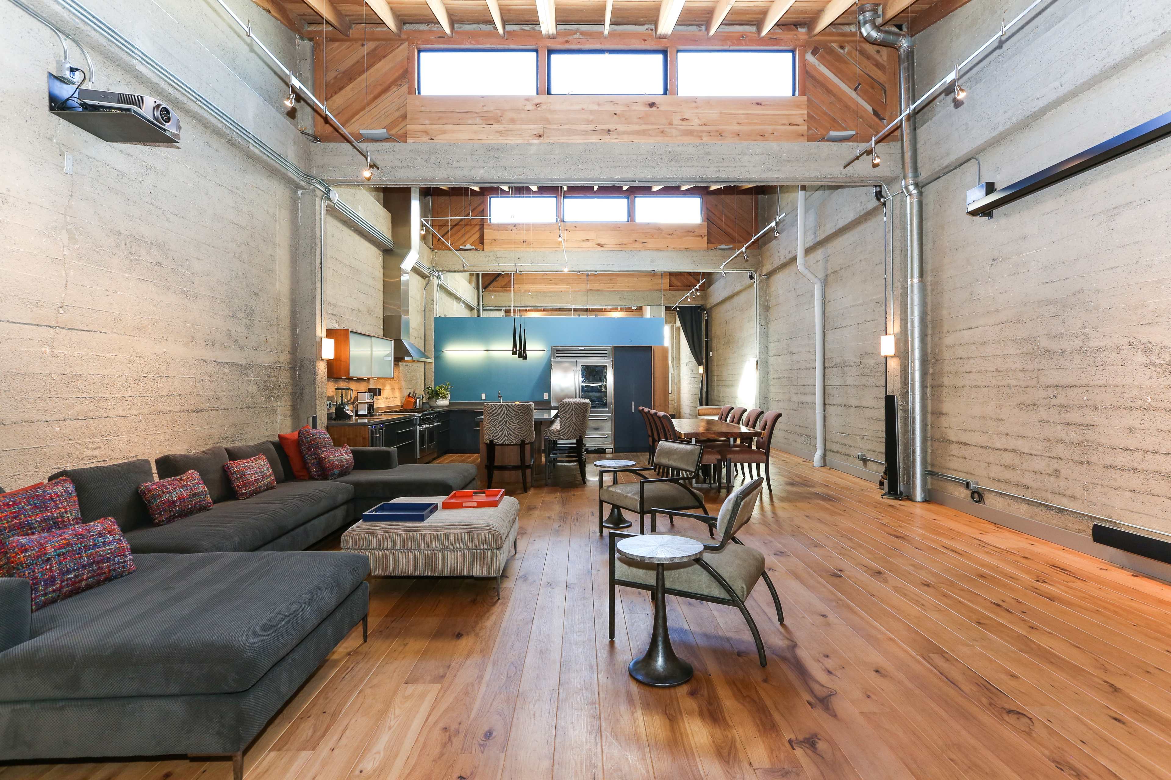 The image shows a spacious, modern living area featuring a sectional sofa, a dining table, and a kitchen at the far end, with wooden beams and concrete walls.