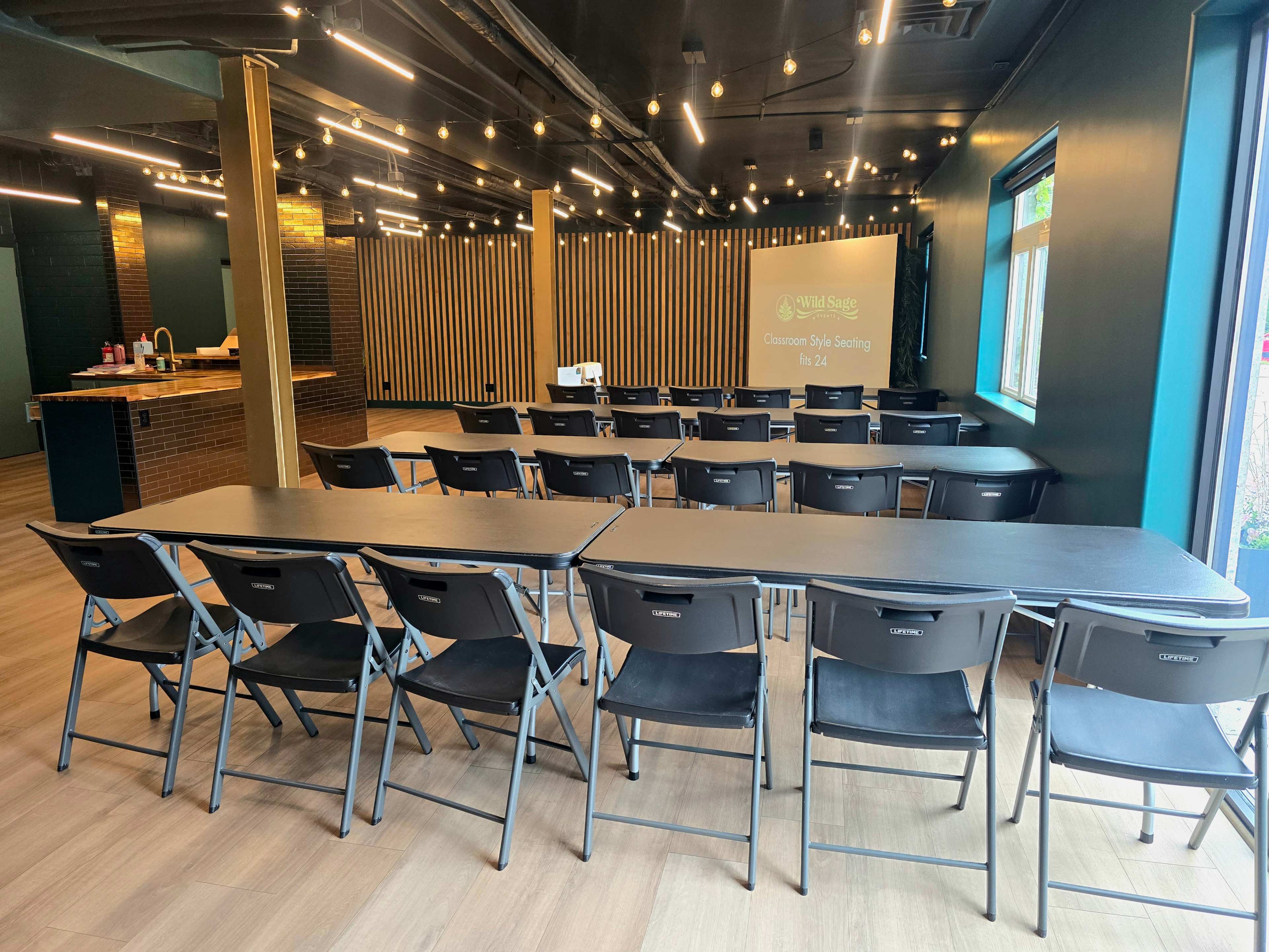 The image depicts a modern classroom setup with rows of folding chairs and tables arranged facing a projector screen in a well-lit room.
