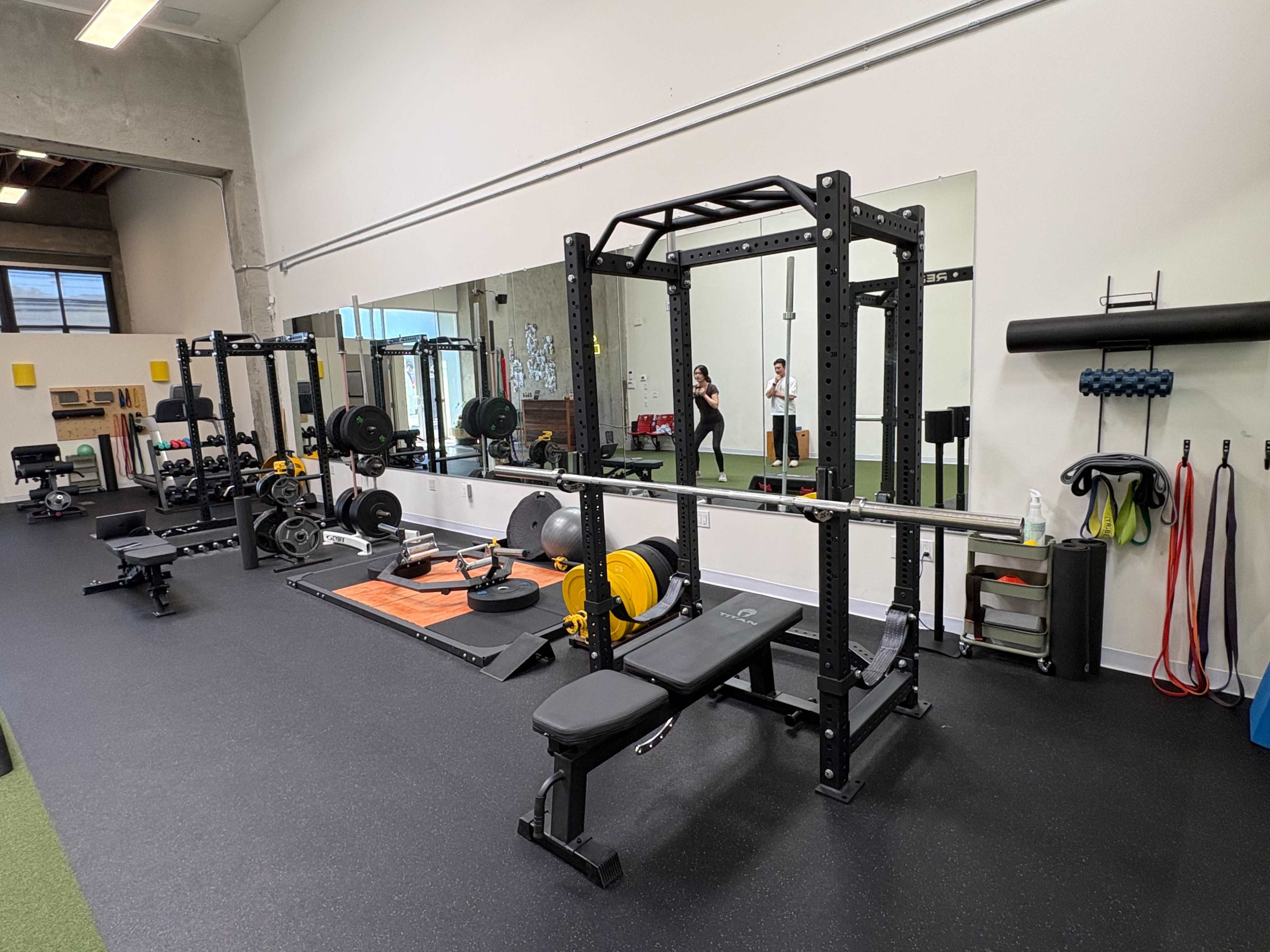 The image shows a gym interior featuring weightlifting equipment, a squat rack, benches, free weights, and a mirrored wall.