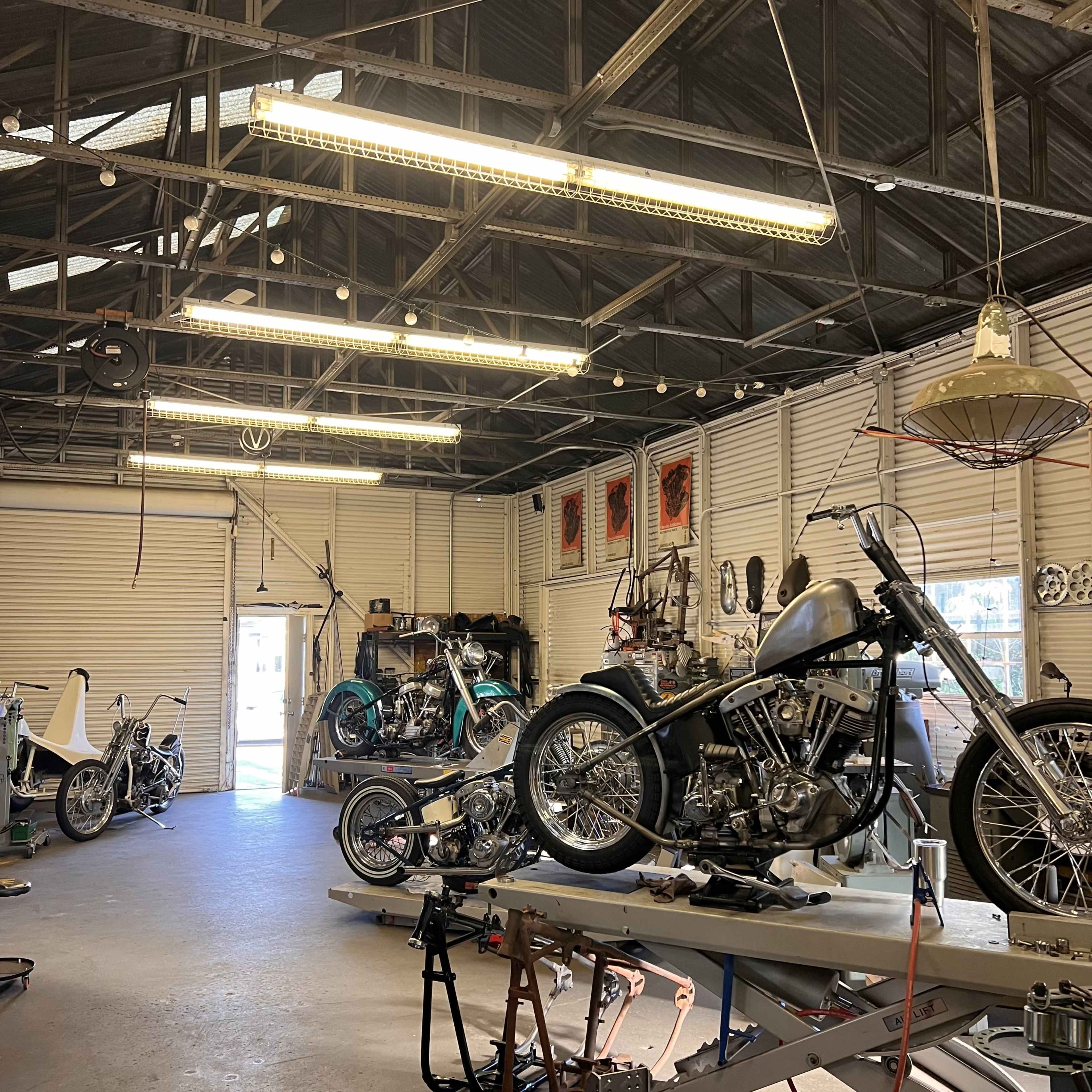 The image shows a workshop filled with several motorcycles in various stages of assembly, along with tools and parts organized on the walls and workbenches.
