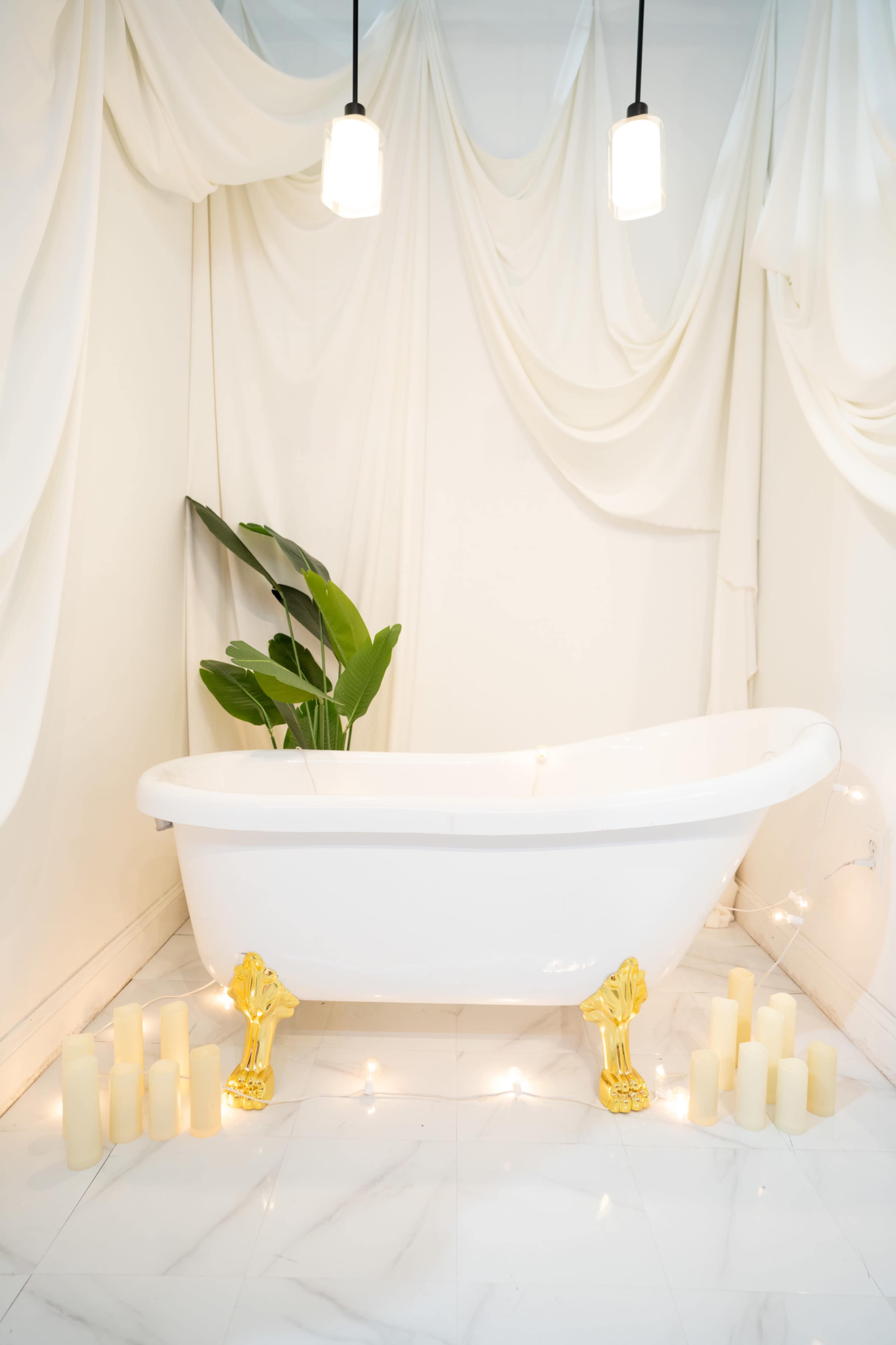 A white clawfoot bathtub with gold legs is positioned in a softly lit room, surrounded by lit candles and a large green plant against a backdrop of flowing white fabric.