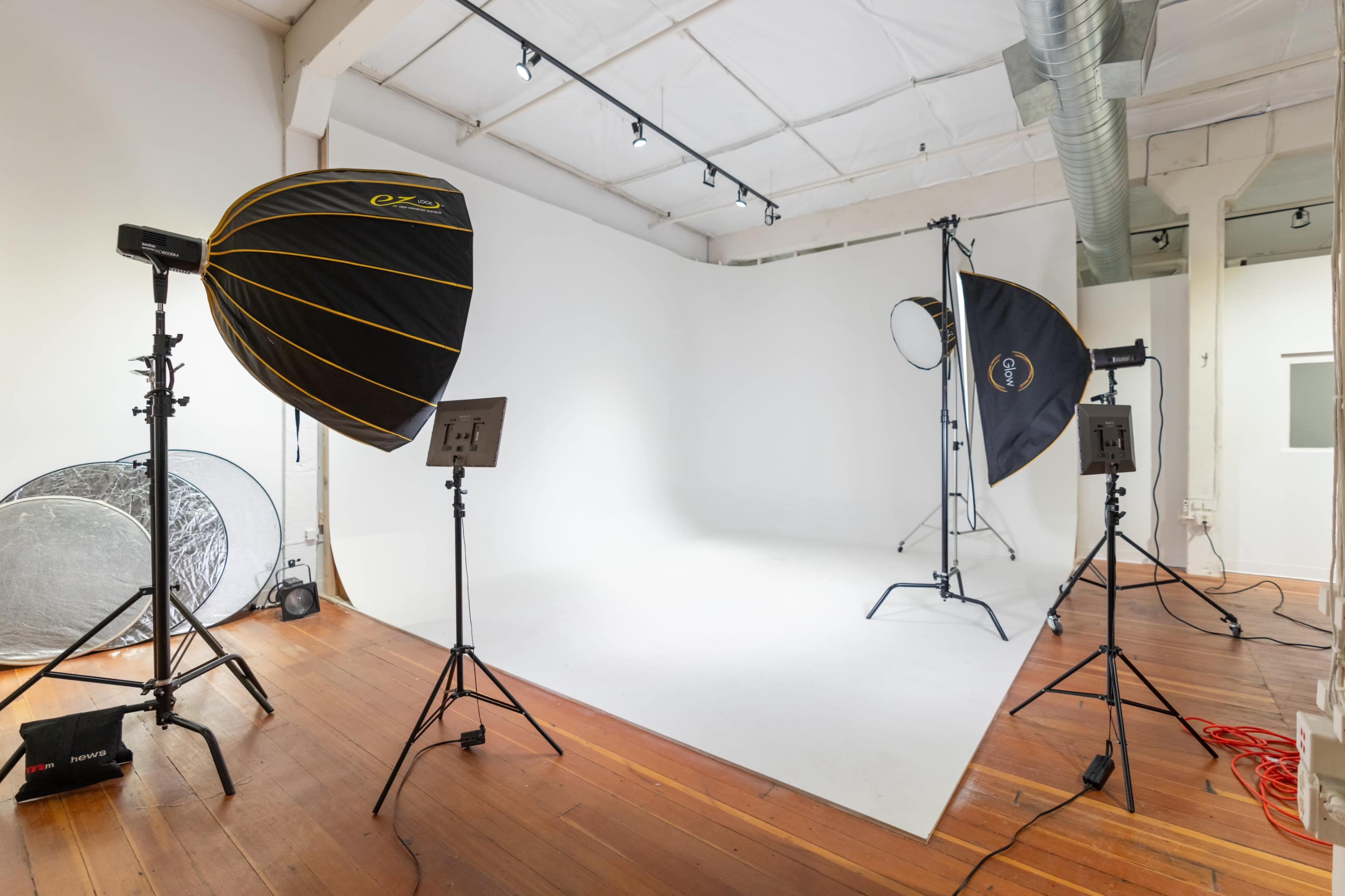 A photography studio with a white backdrop, two softbox lights, a reflector, and wooden flooring.