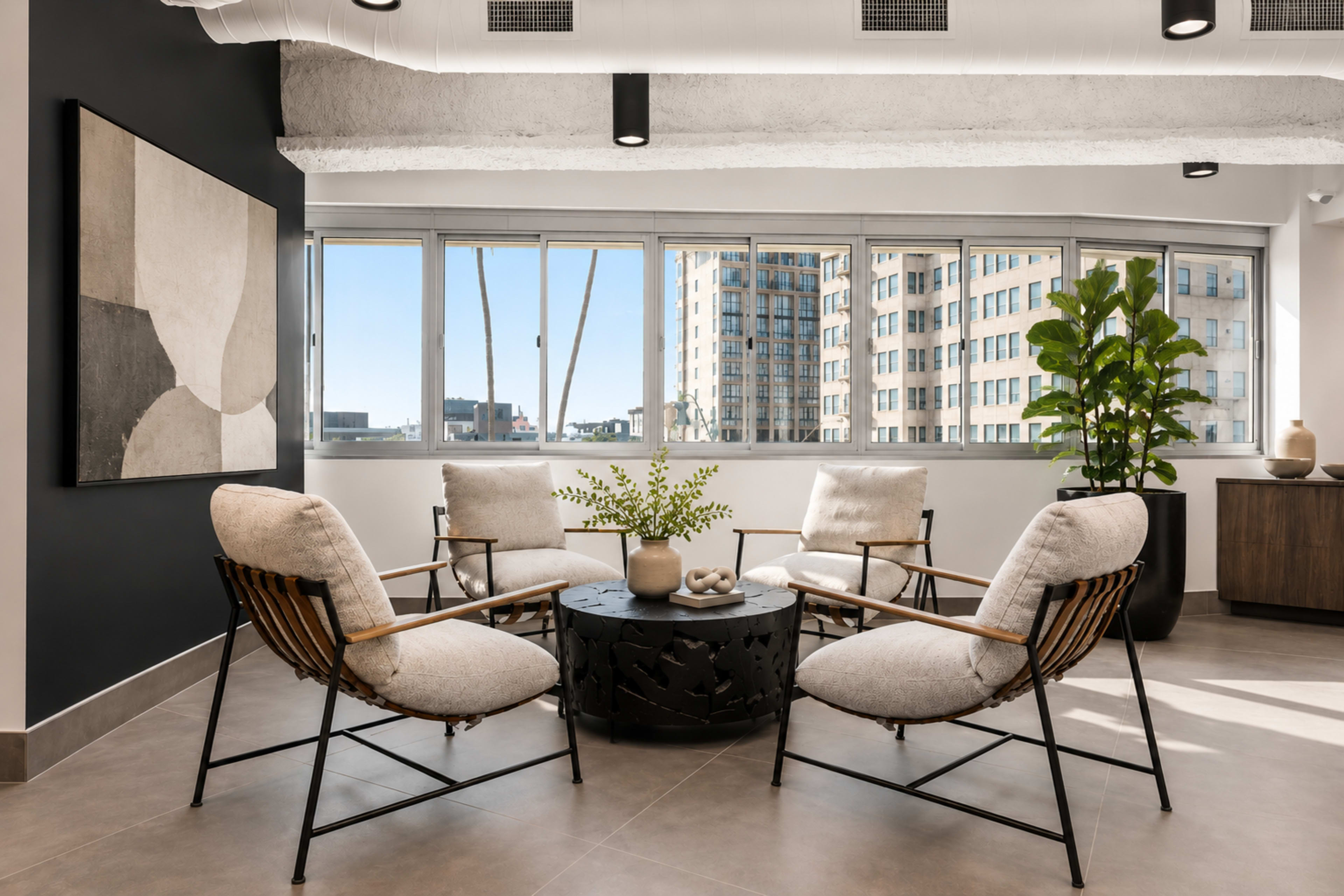A modern seating area features four upholstered chairs arranged around a circular table, with large windows providing a view of the city skyline and a potted plant in the corner.