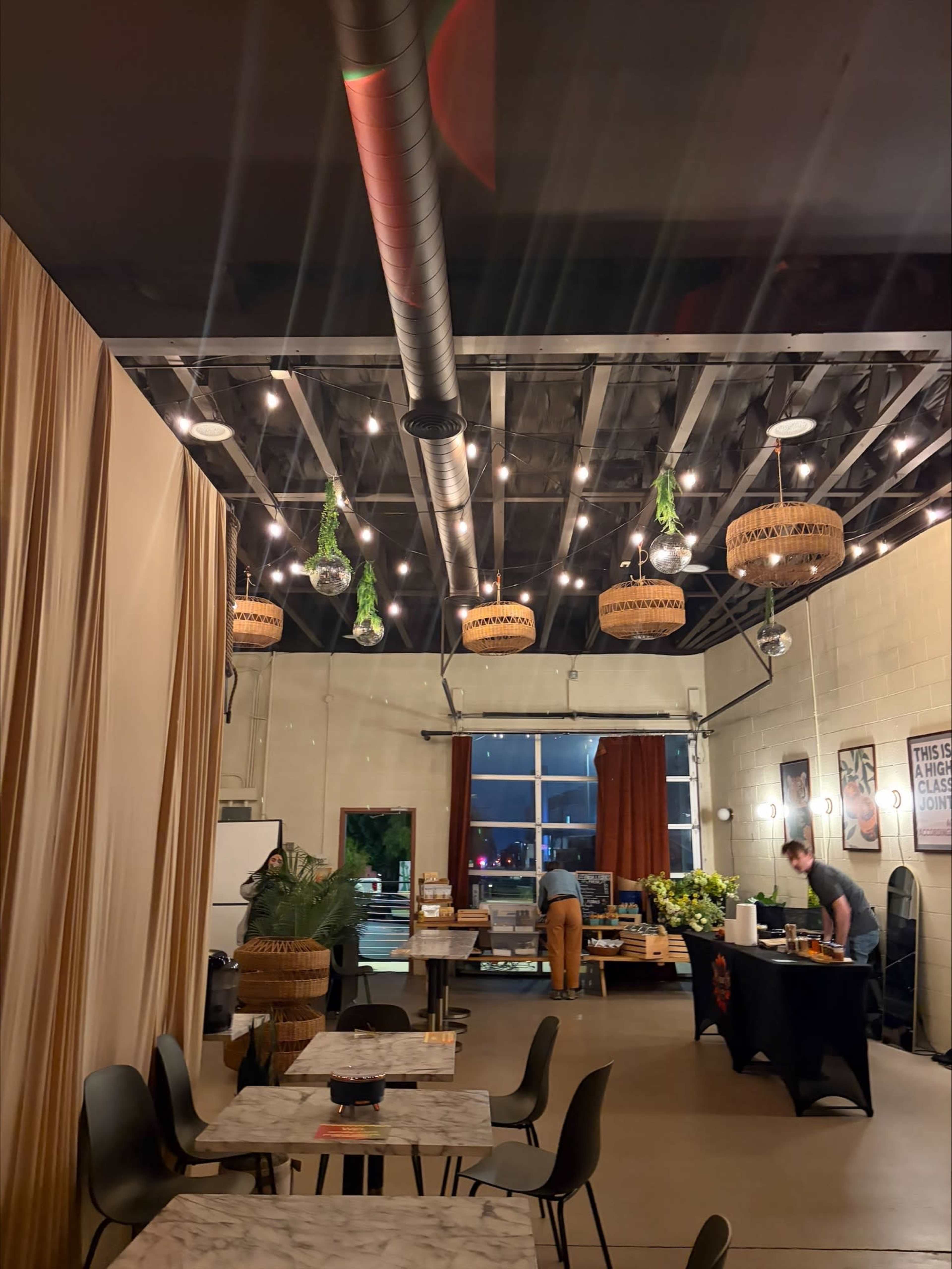 The image shows a modern café interior with exposed beams, hanging pendant lights, and a table setup in the foreground, while a staff member organizes items in the background.