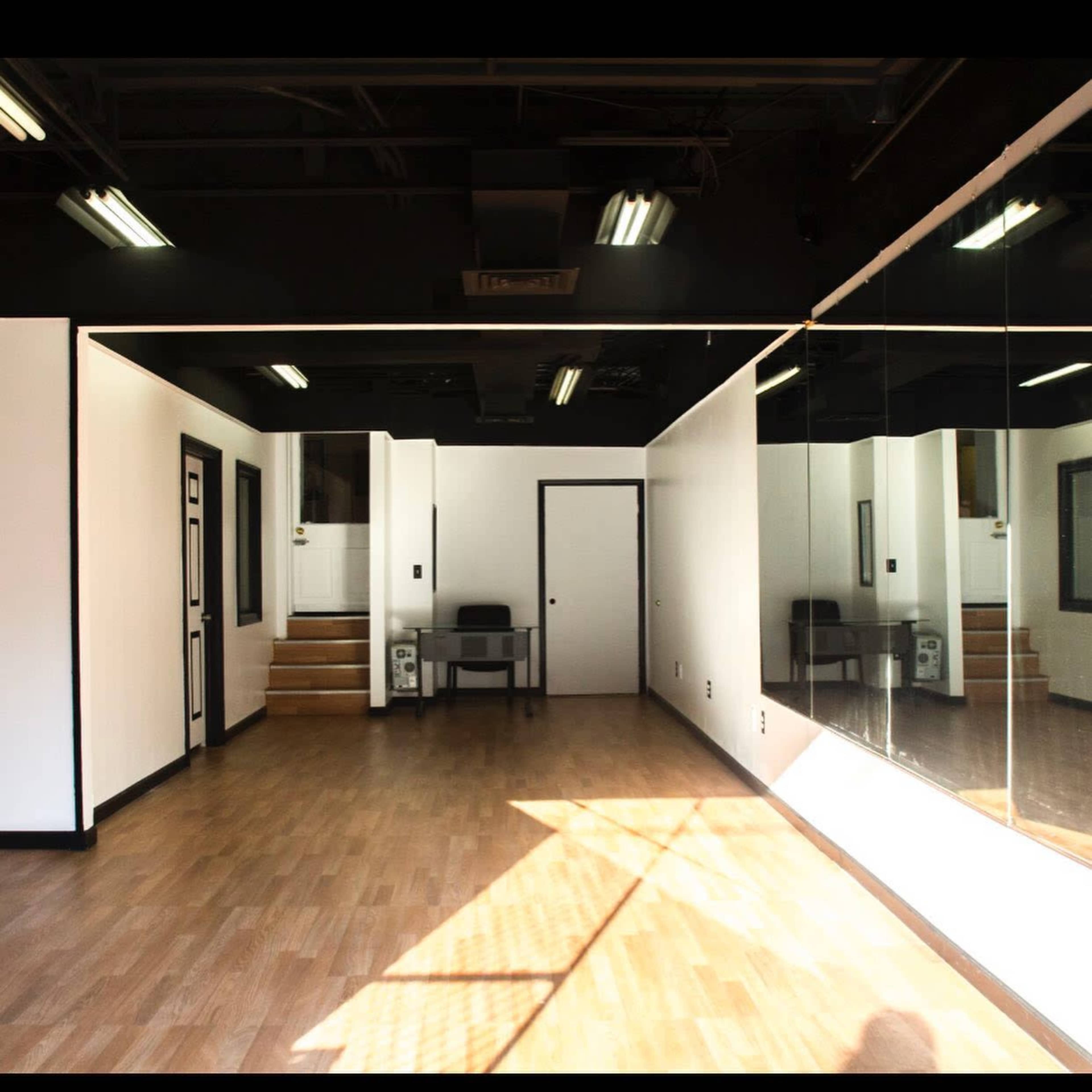 The image shows a spacious room with wooden flooring, a large mirror on one wall, a small table and chair, and a staircase in the background.