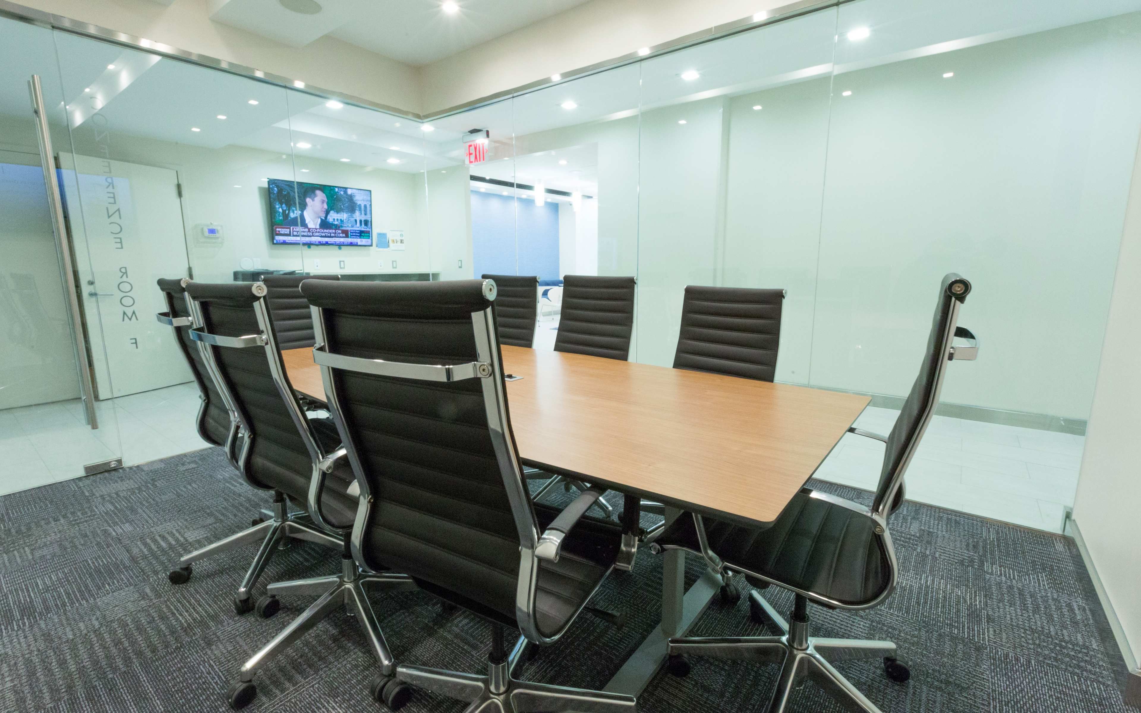 A modern conference room features a large wooden table surrounded by black ergonomic chairs, with a TV mounted on the wall displaying news.