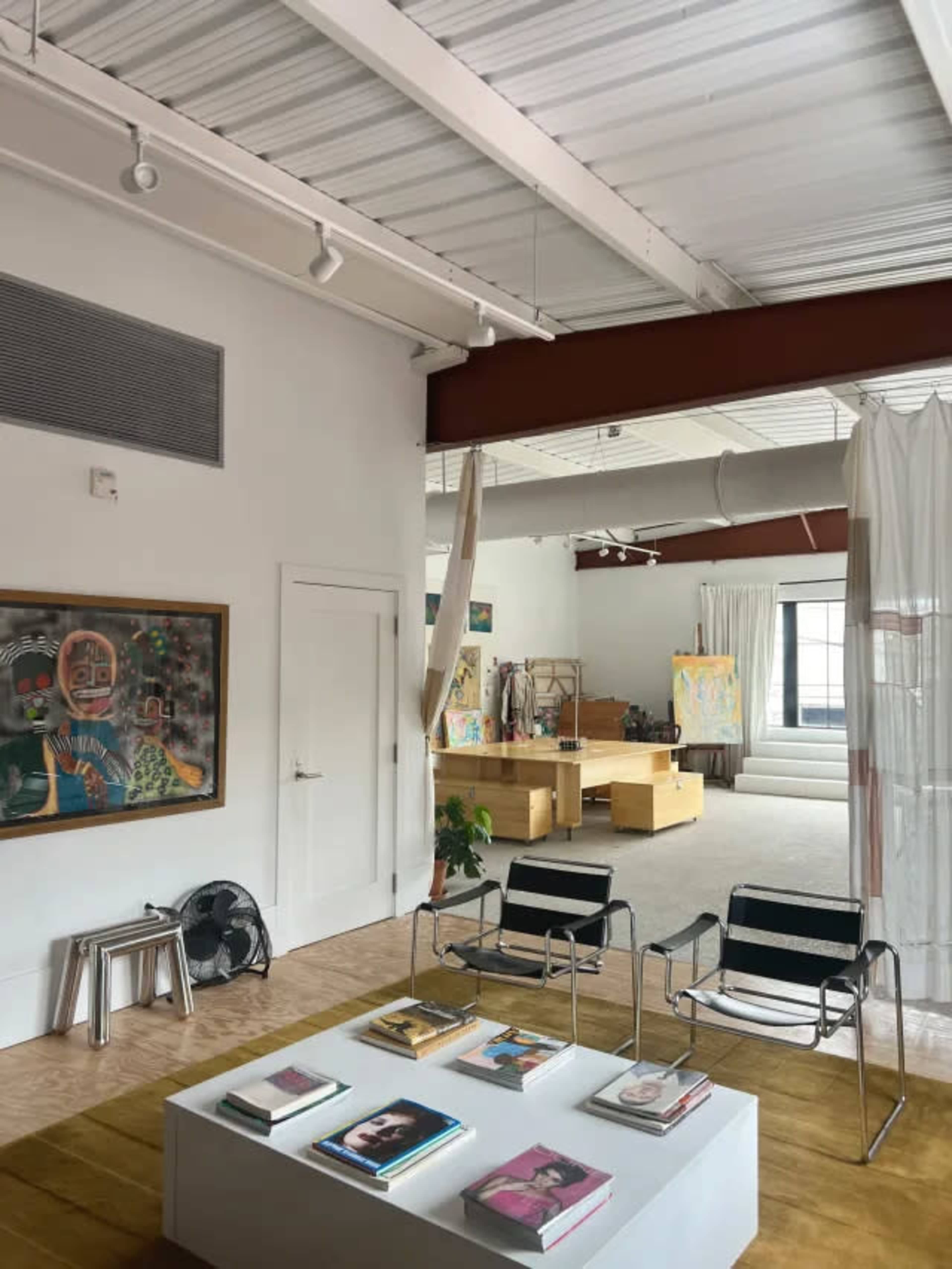 The image shows a modern indoor space featuring a seating area with two chairs, a coffee table stacked with books, and open walls leading to a brightly lit studio area.