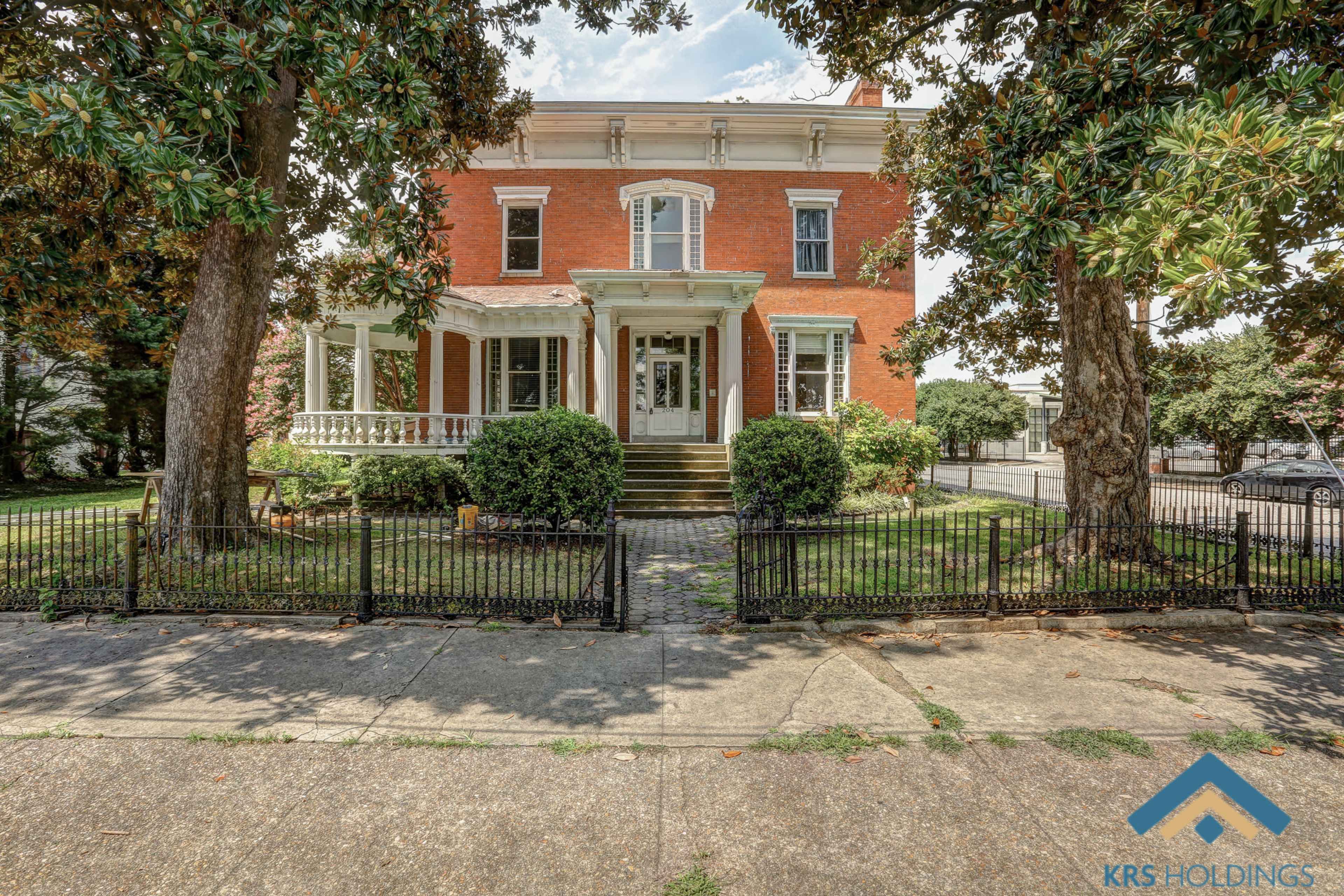 A classic two-story red brick house with a white front porch and a well-maintained garden, enclosed by a black wrought iron fence.