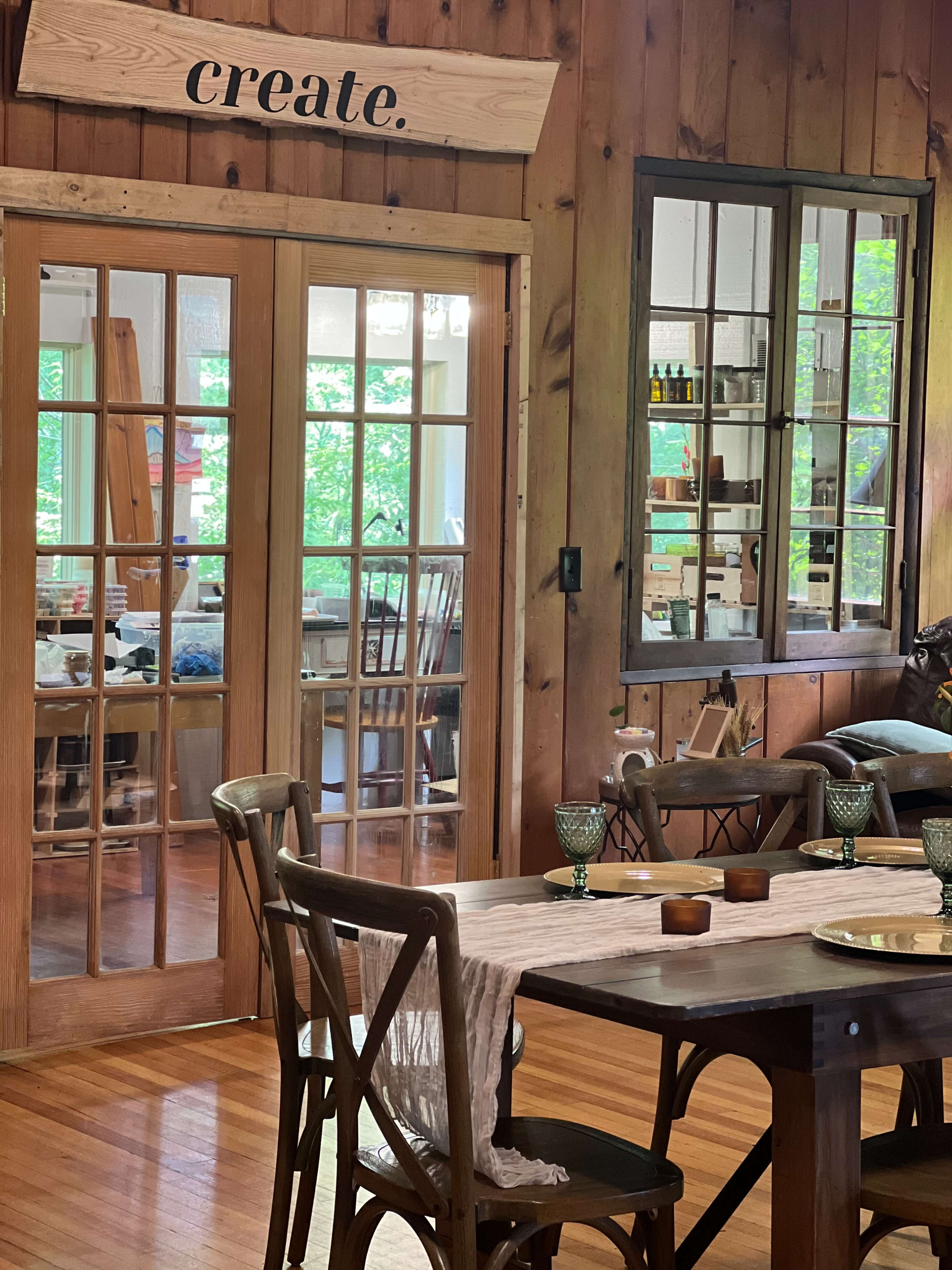 A wooden-floored room features a dining table set with decorative items, leading to a bright workshop through glass double doors.