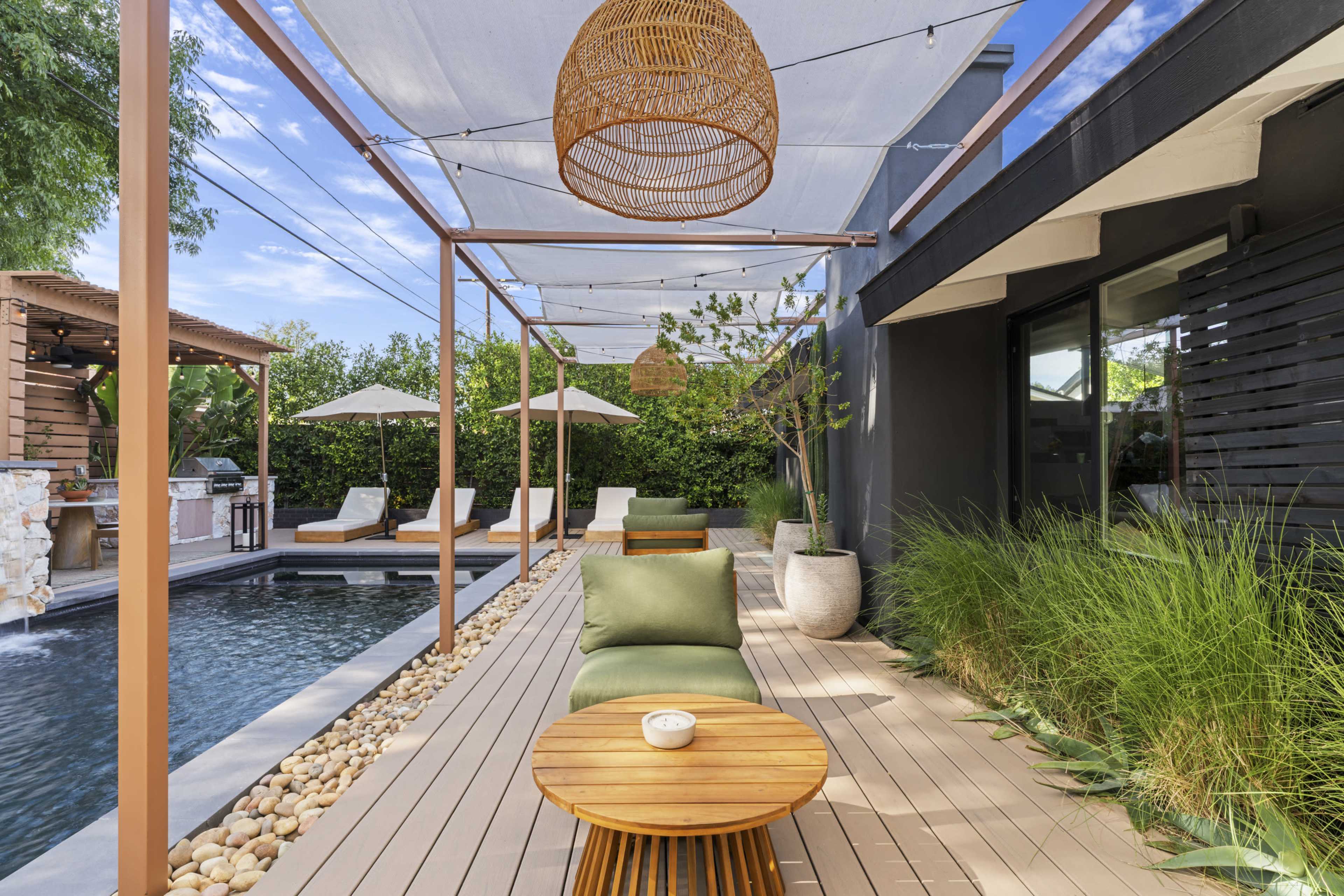 A modern outdoor space features a swimming pool, lounge chairs, a small wooden table, and a shaded seating area with decorative plants.