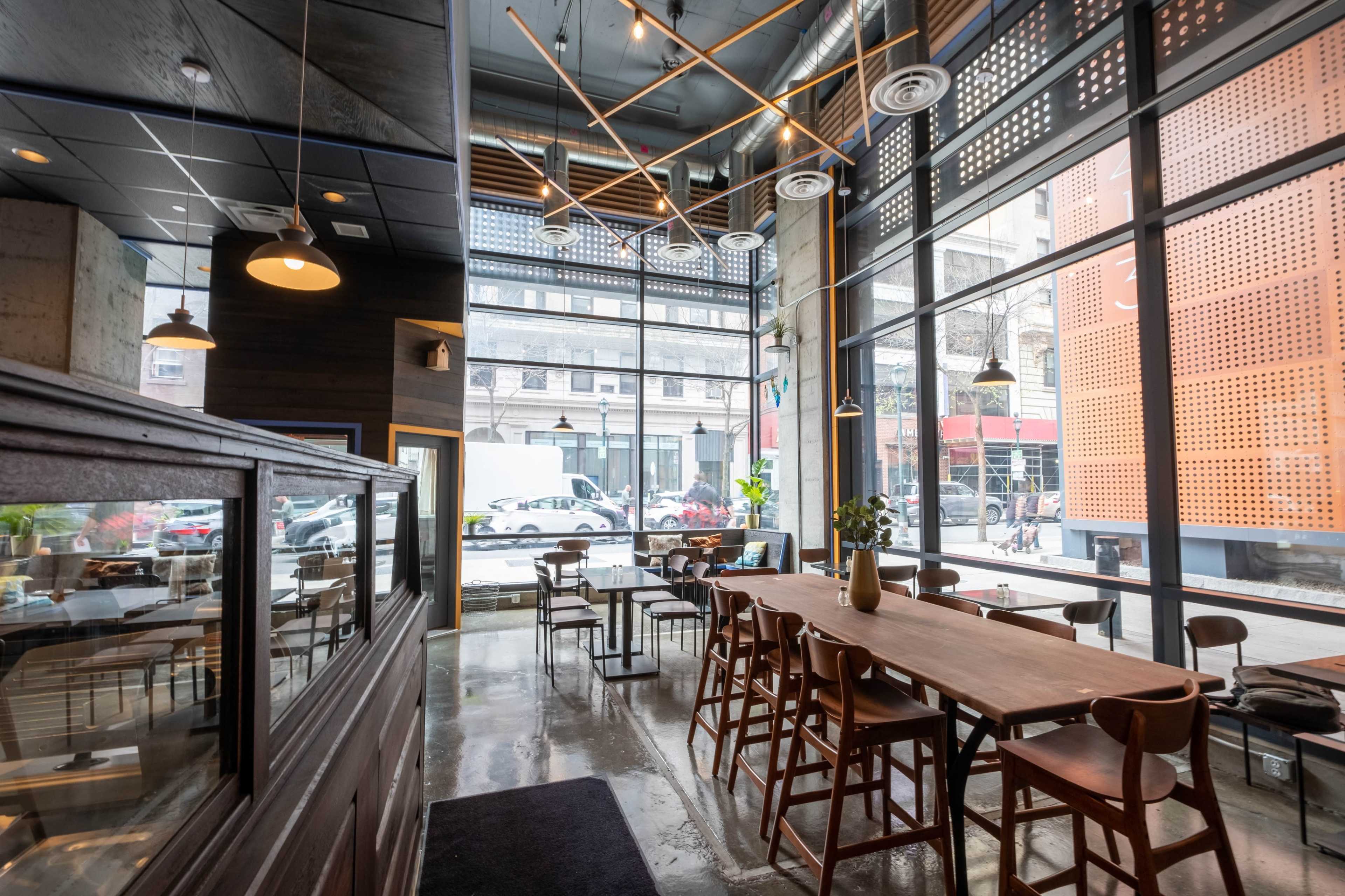 A modern cafe interior featuring wooden tables, chairs, and large windows with a view of the street outside.