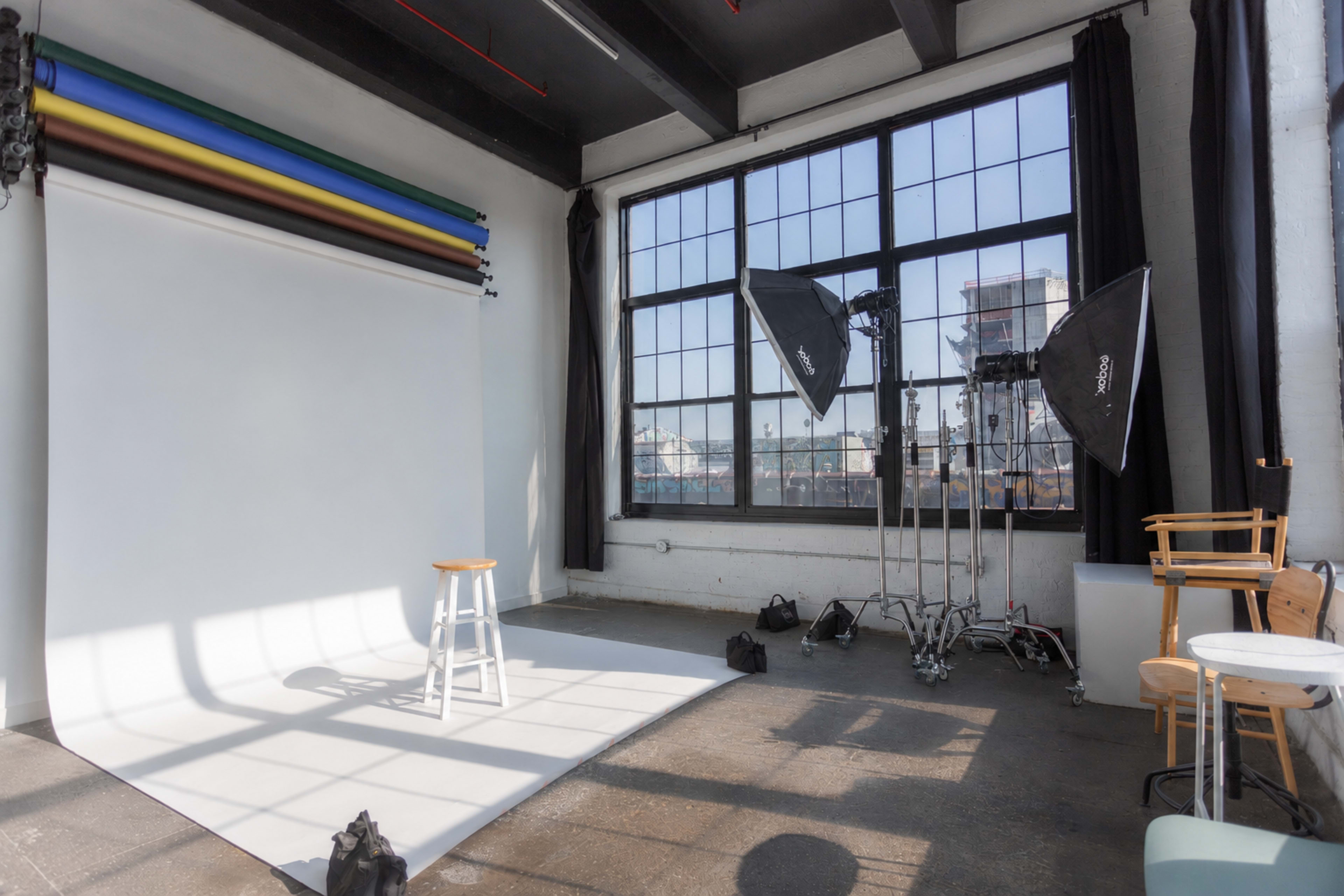 The image shows a photography studio with a white backdrop, a stool in the center, and large softbox lights positioned nearby.