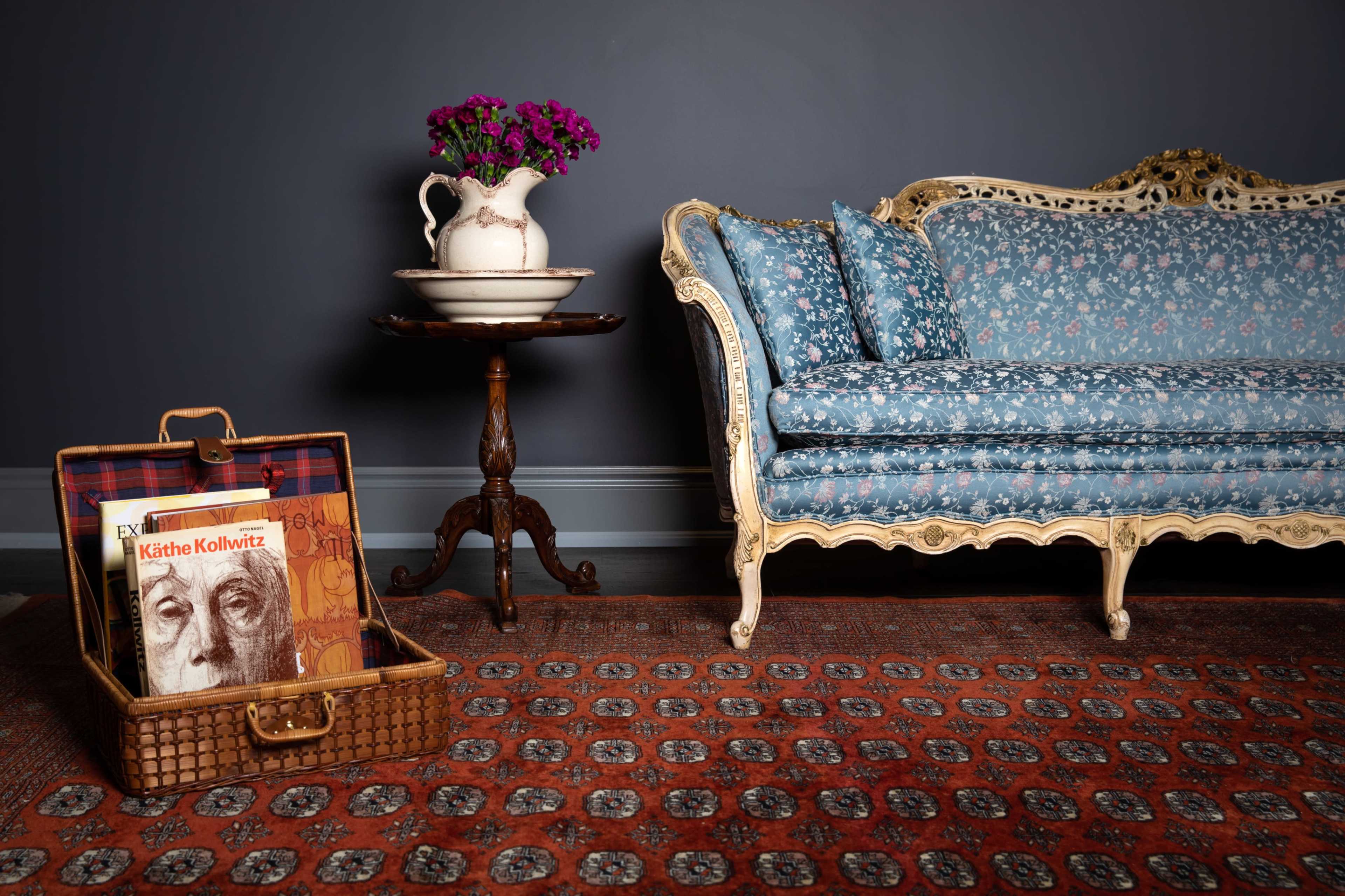 A vintage loveseat with floral upholstery is positioned next to a small table featuring a vase of flowers and a wicker suitcase containing books.