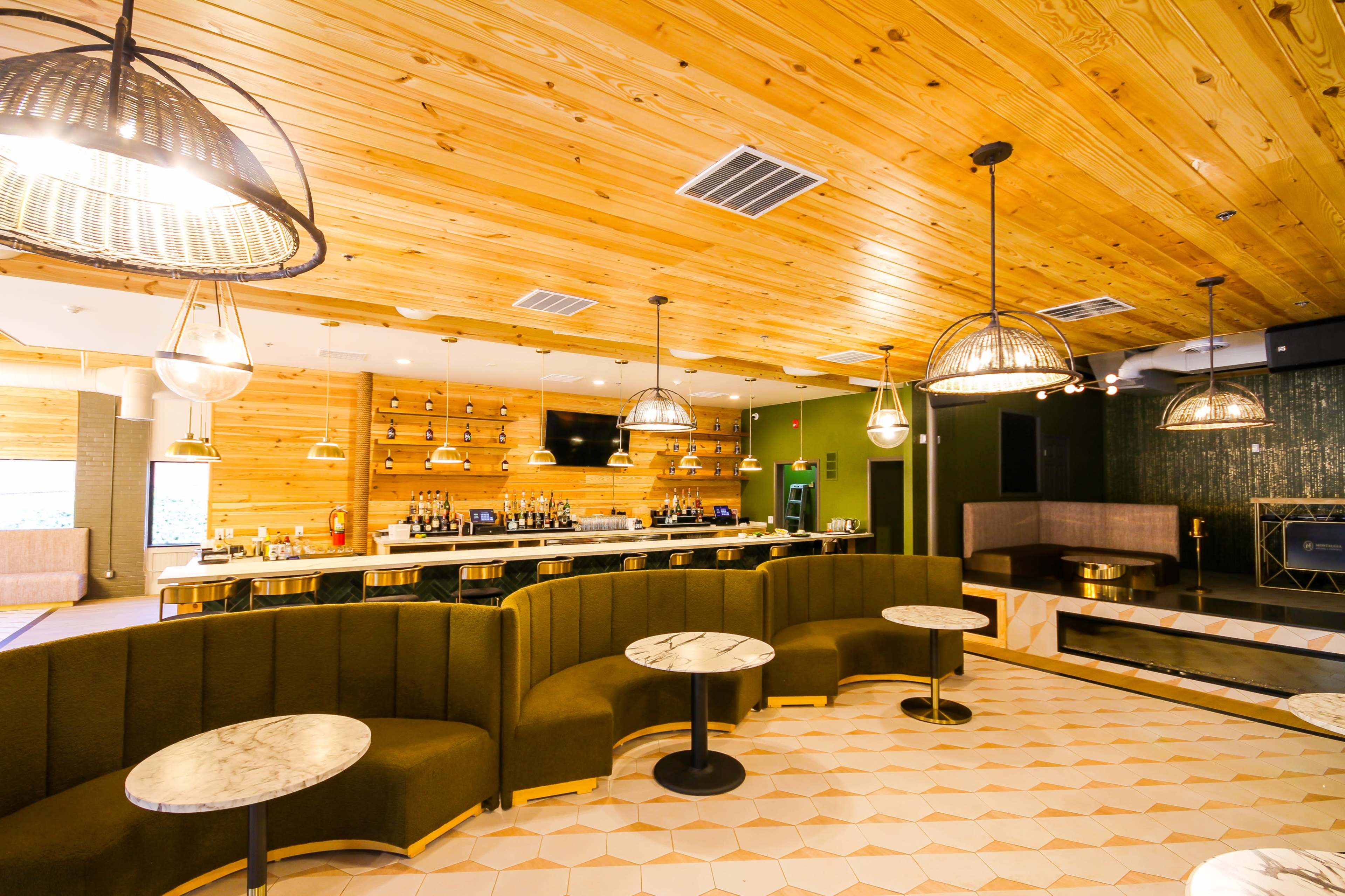 A modern lounge area featuring wooden walls and a ceiling, with a curved green seating section and marble-topped tables under decorative pendant lights.