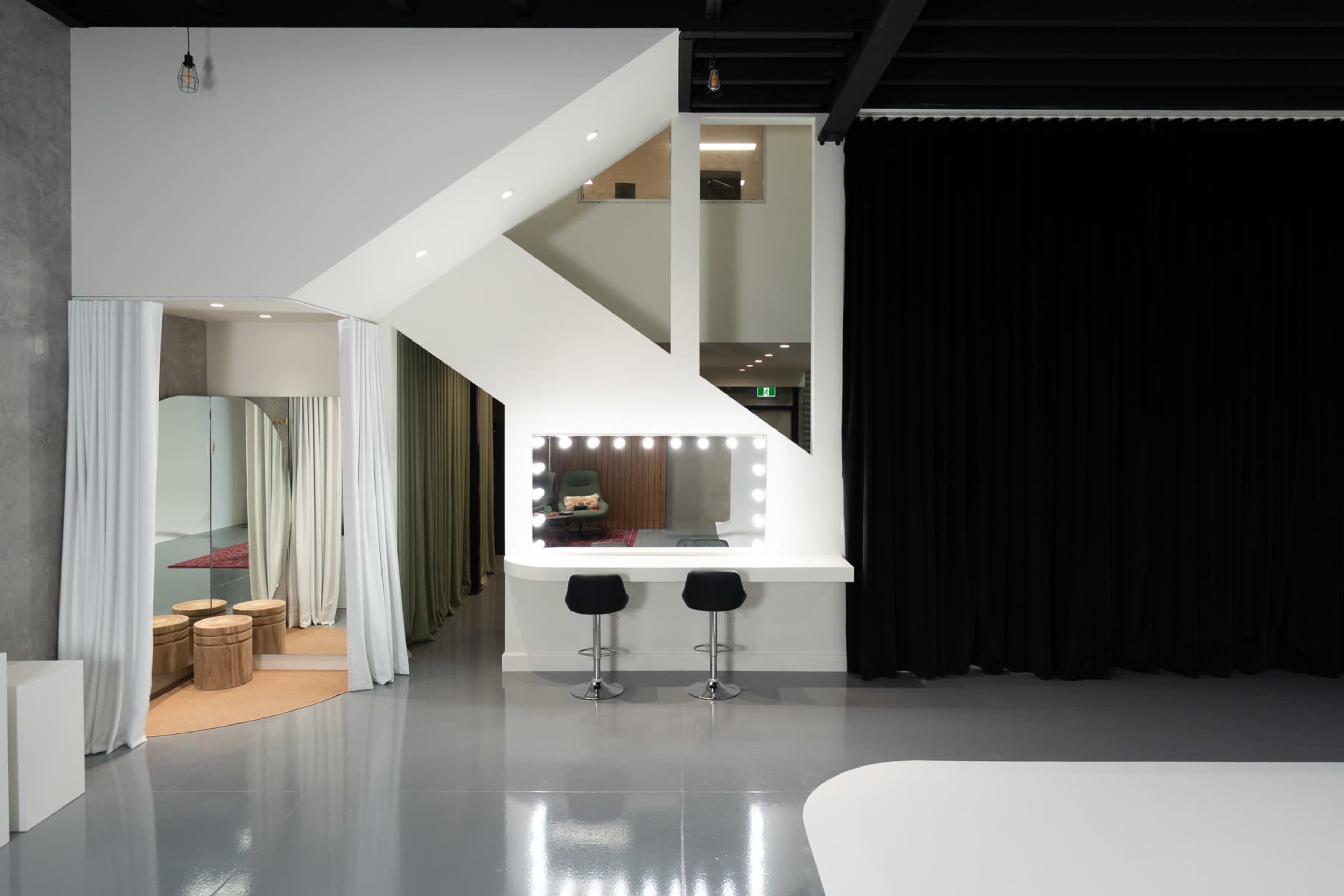 The image shows a modern studio space featuring a staircase, a vanity with lit mirrors, and seating, alongside a black curtain and light-colored flooring.