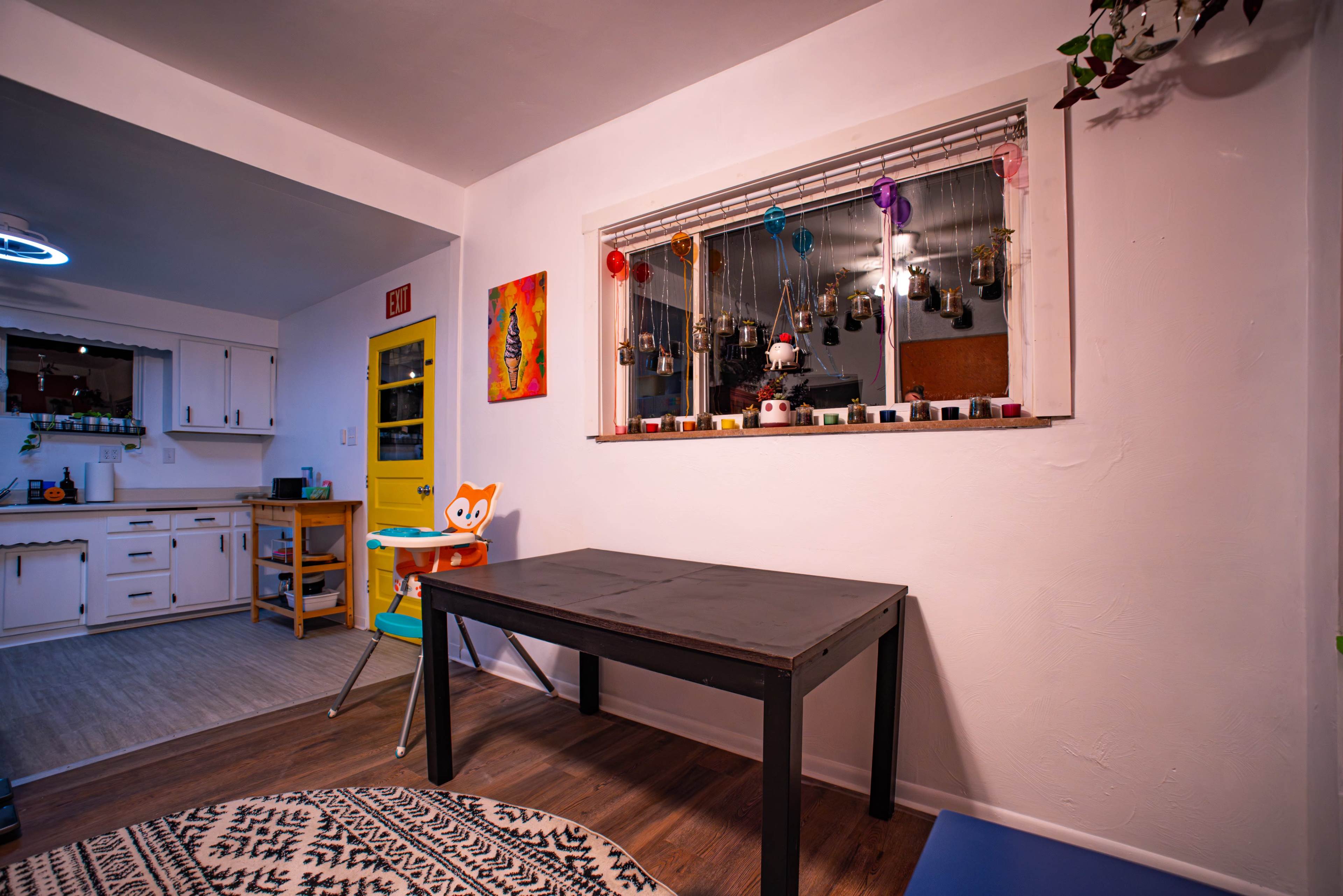 A black table sits in a room with a window displaying colorful hanging decor, while a kitchen area is visible in the background.