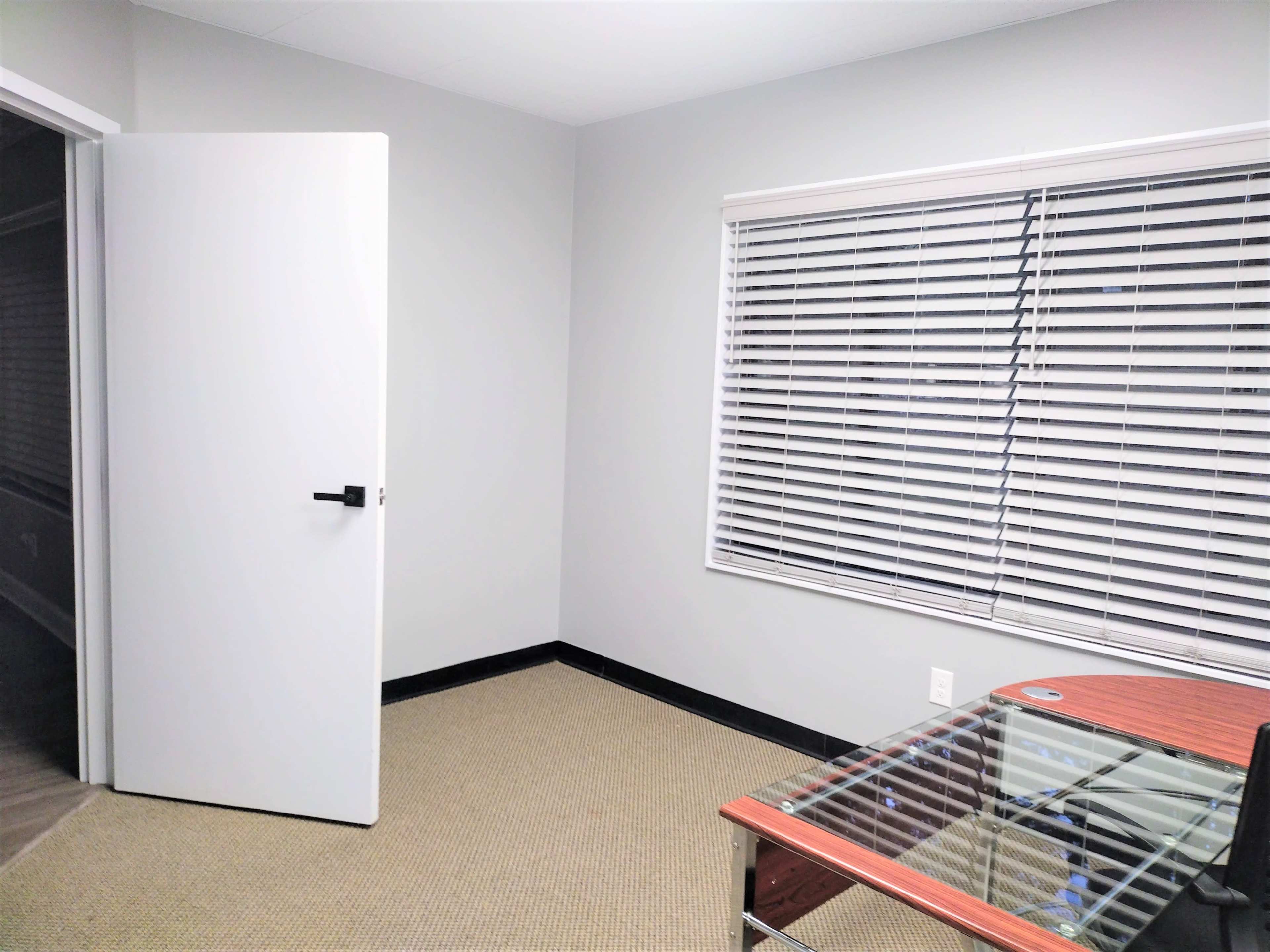 A bare room with a glass-top desk, a door ajar, and a window with closed blinds.