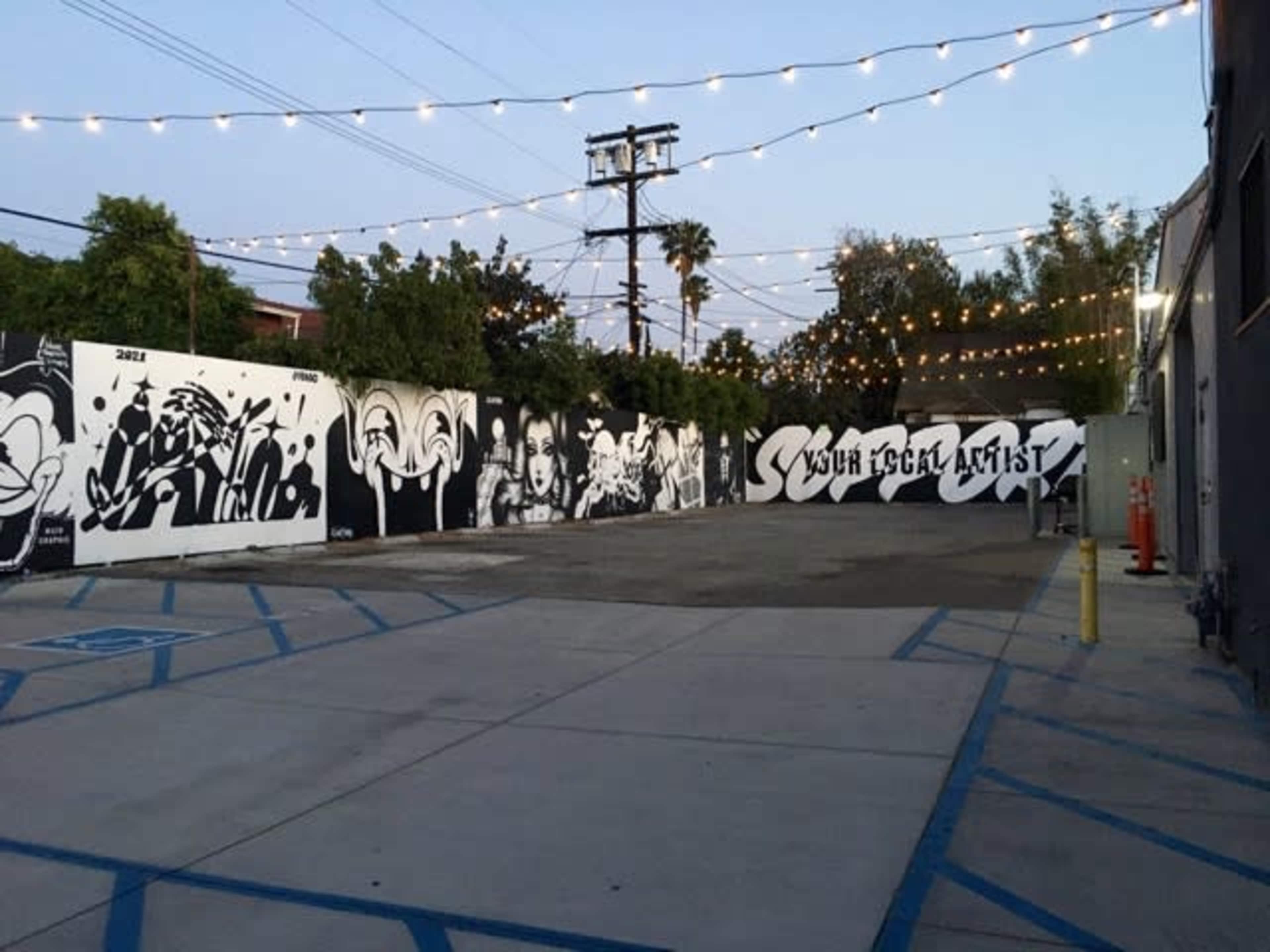 The image shows an outdoor mural-covered area with string lights above, featuring several large black and white artworks on the walls.