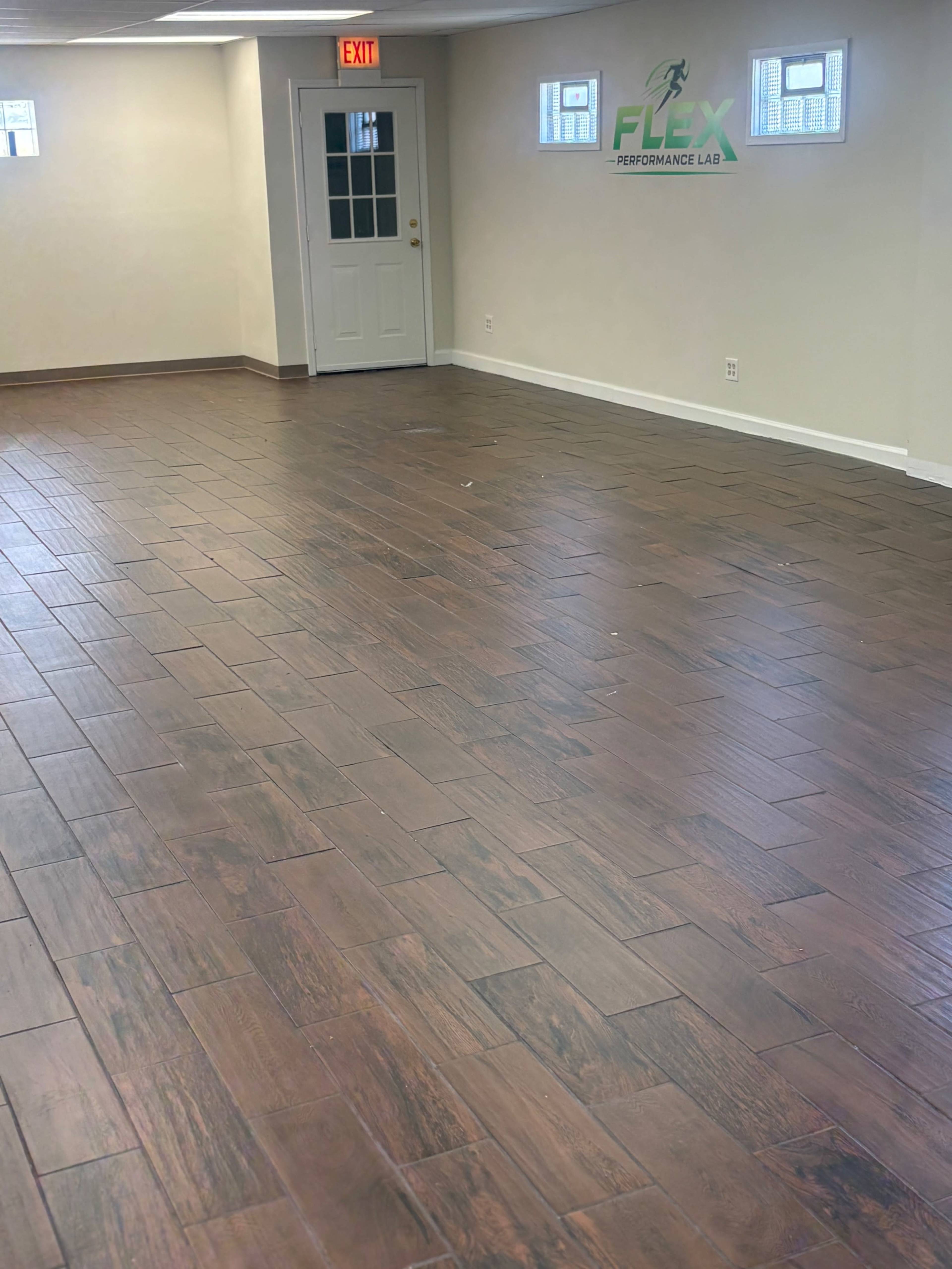 The image shows an empty room with wooden-style tiled flooring, a closed door, and a wall sign that reads "FLEX PERFORMANCE LAB."