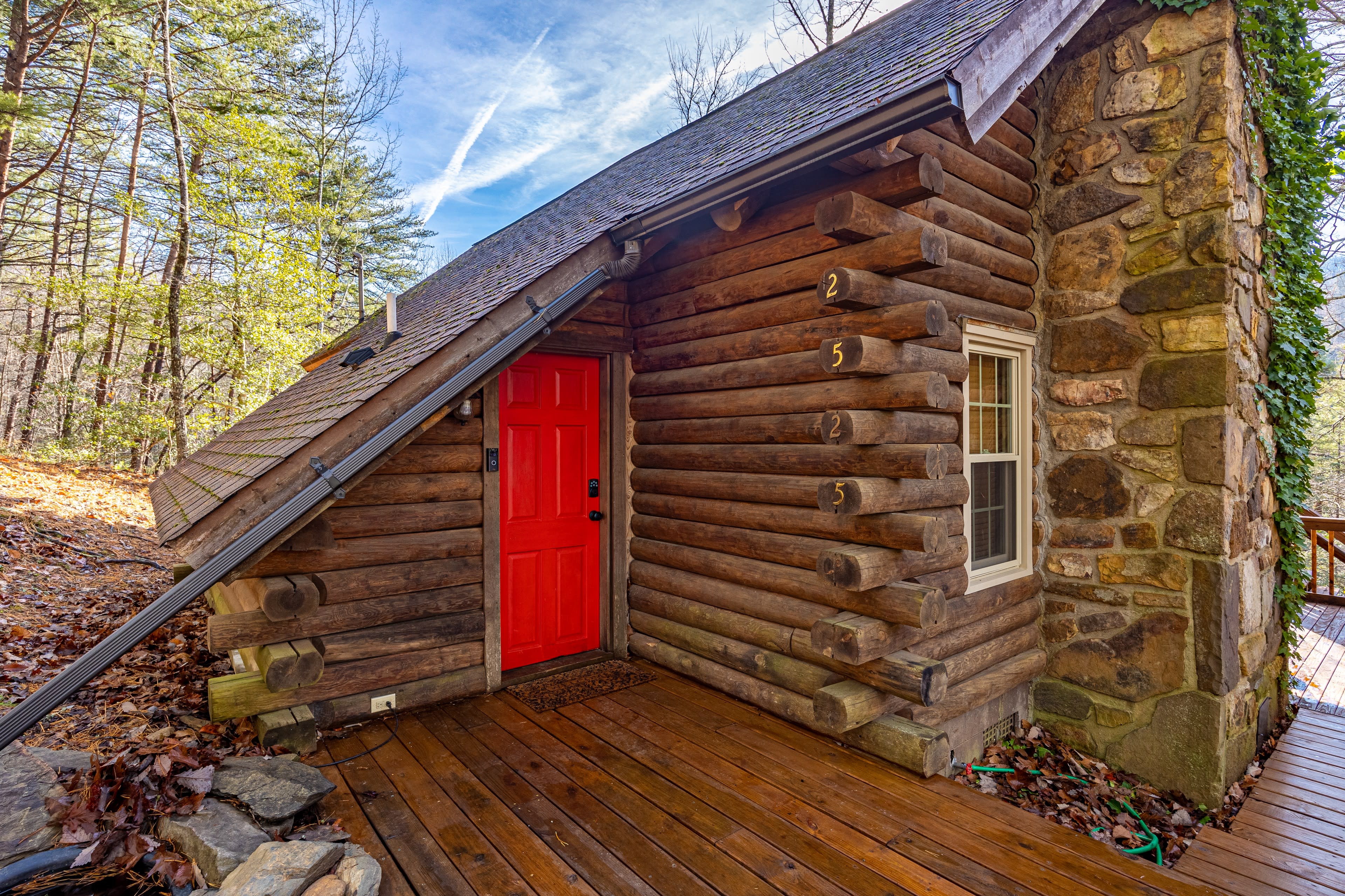 A log cabin with a bright red door stands amidst a wooded area, featuring a stone wall and wooden deck.