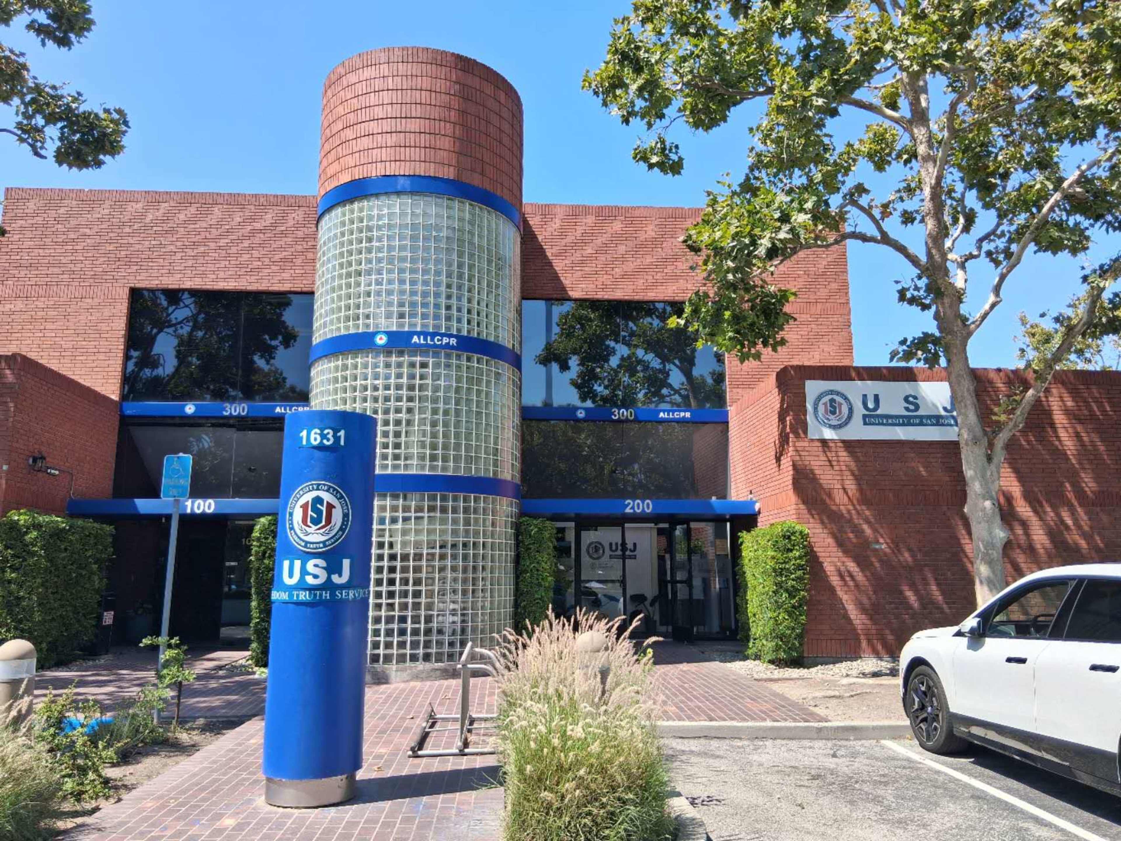 The image shows a modern two-story building with a glass and brick exterior labeled "USJ," set among trees and landscaping.
