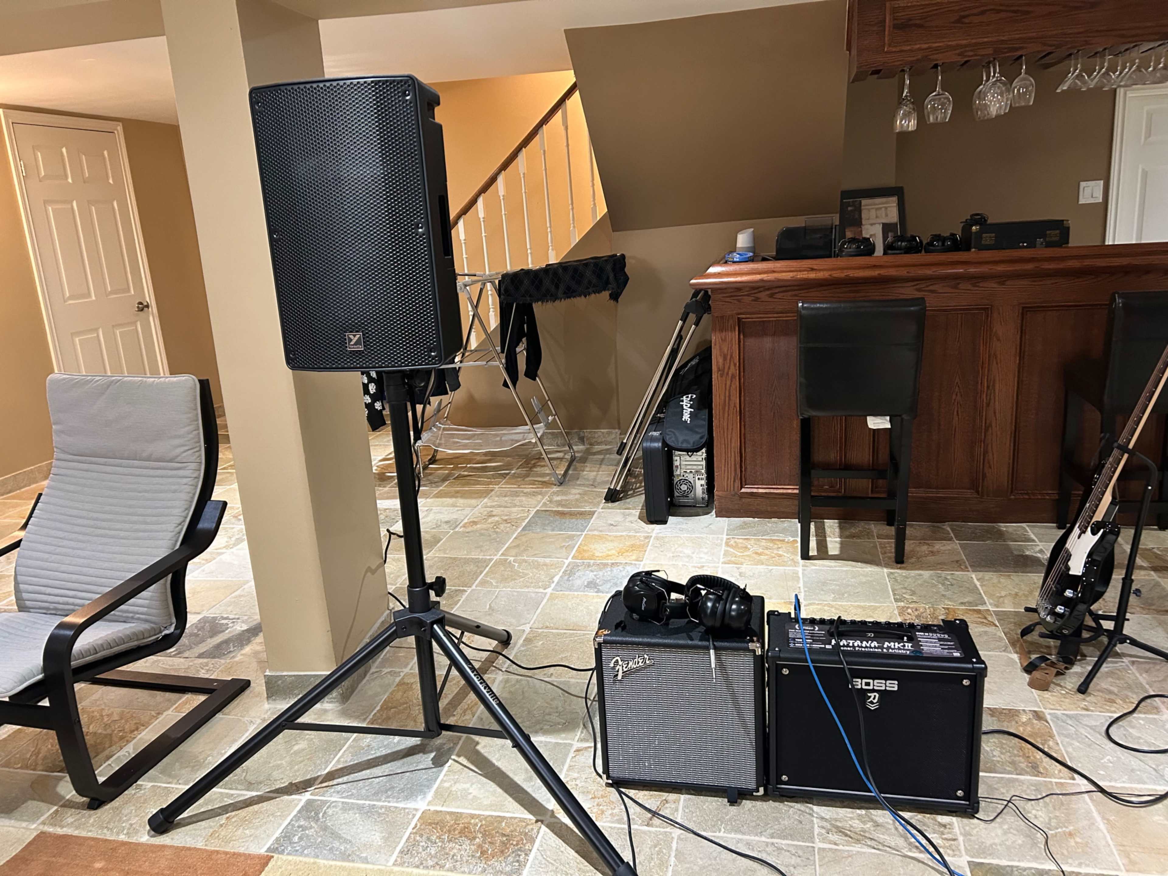 A speaker on a stand next to two amplifiers and headphones, with a bar area and a folding chair in the background.