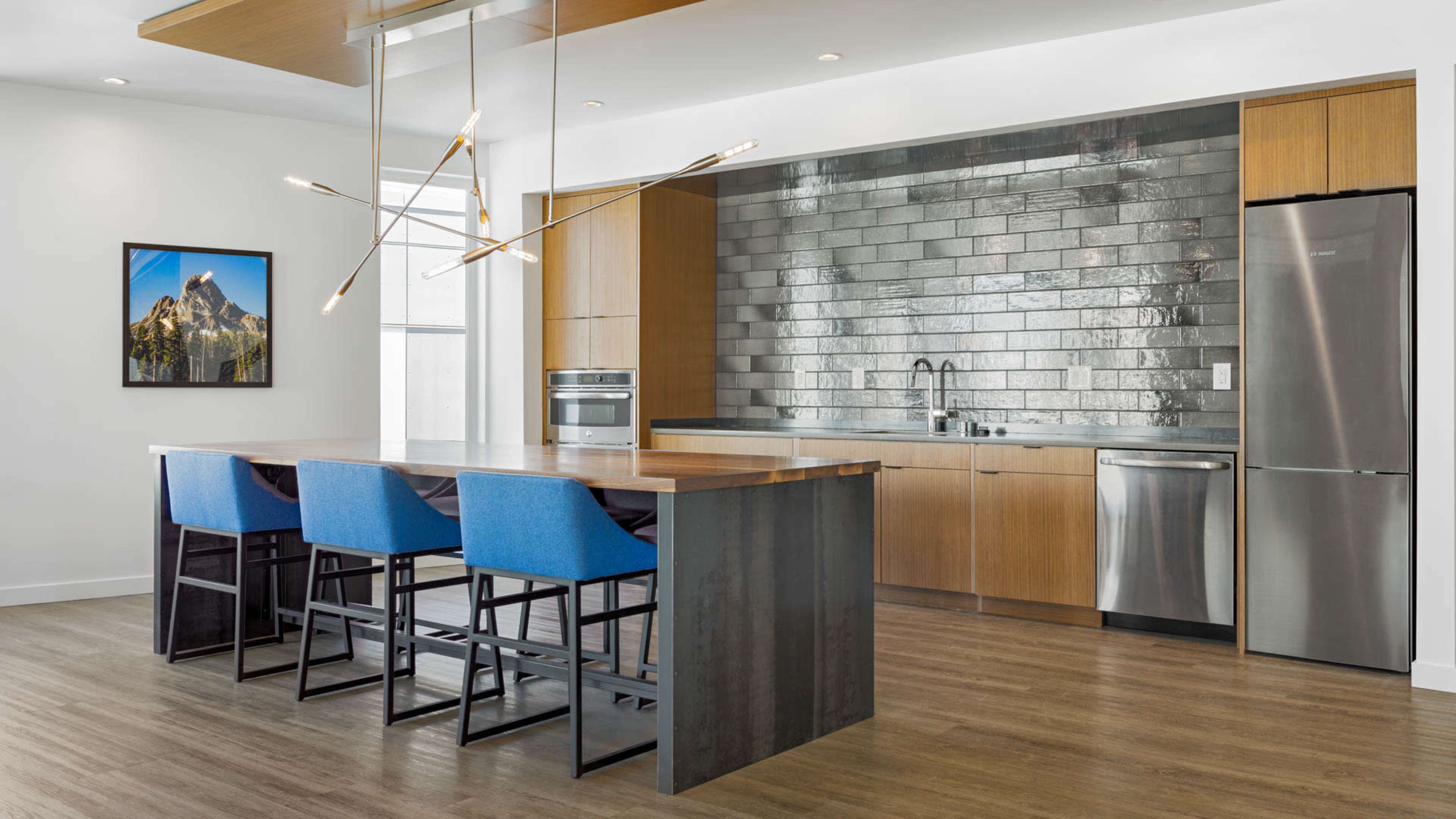 A modern kitchen with a long wooden island, blue bar stools, stainless steel appliances, and a tiled backsplash.