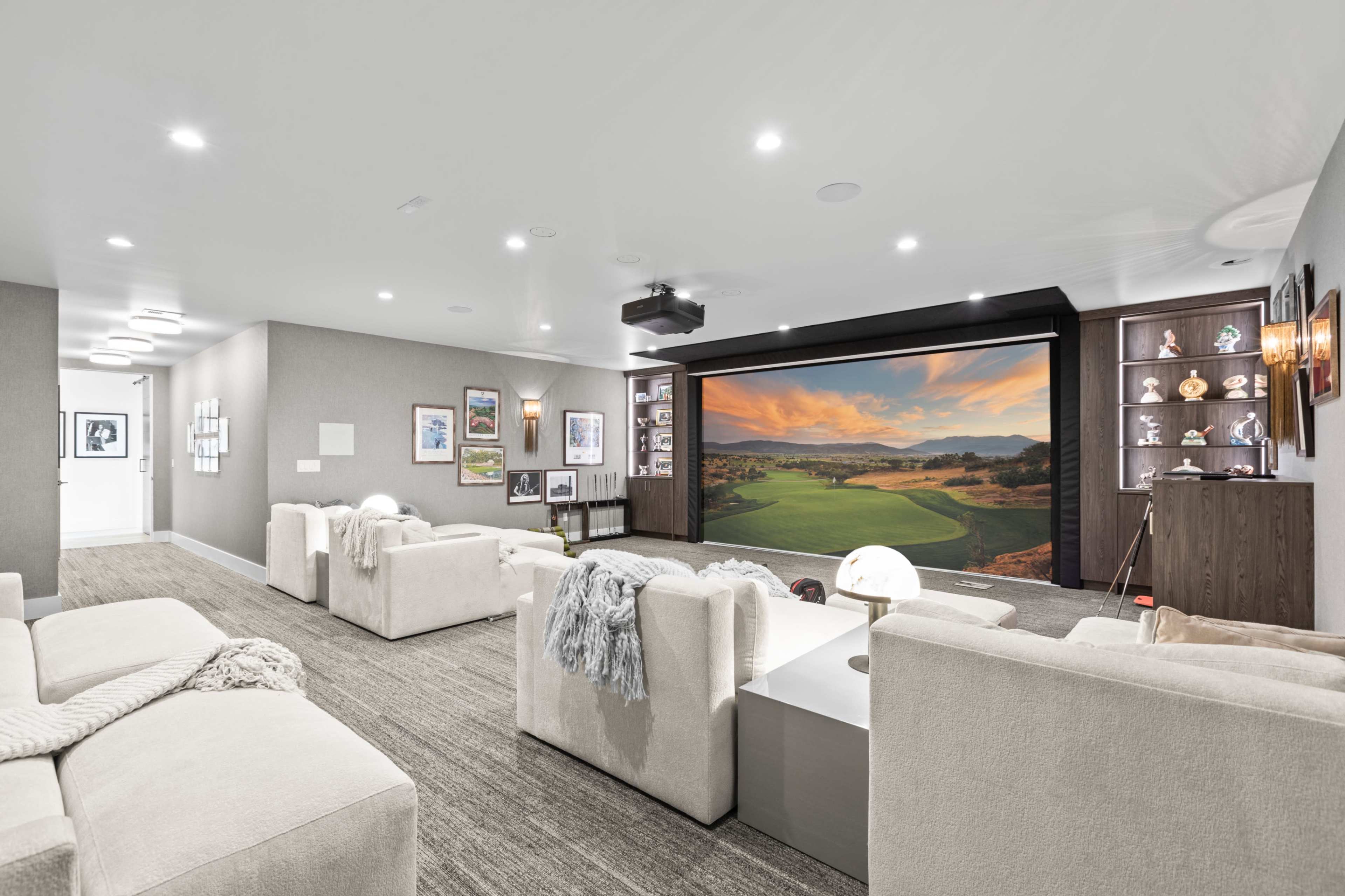 A modern home theater with white sofas, a large projection screen displaying a landscape, and various framed pictures on the walls.