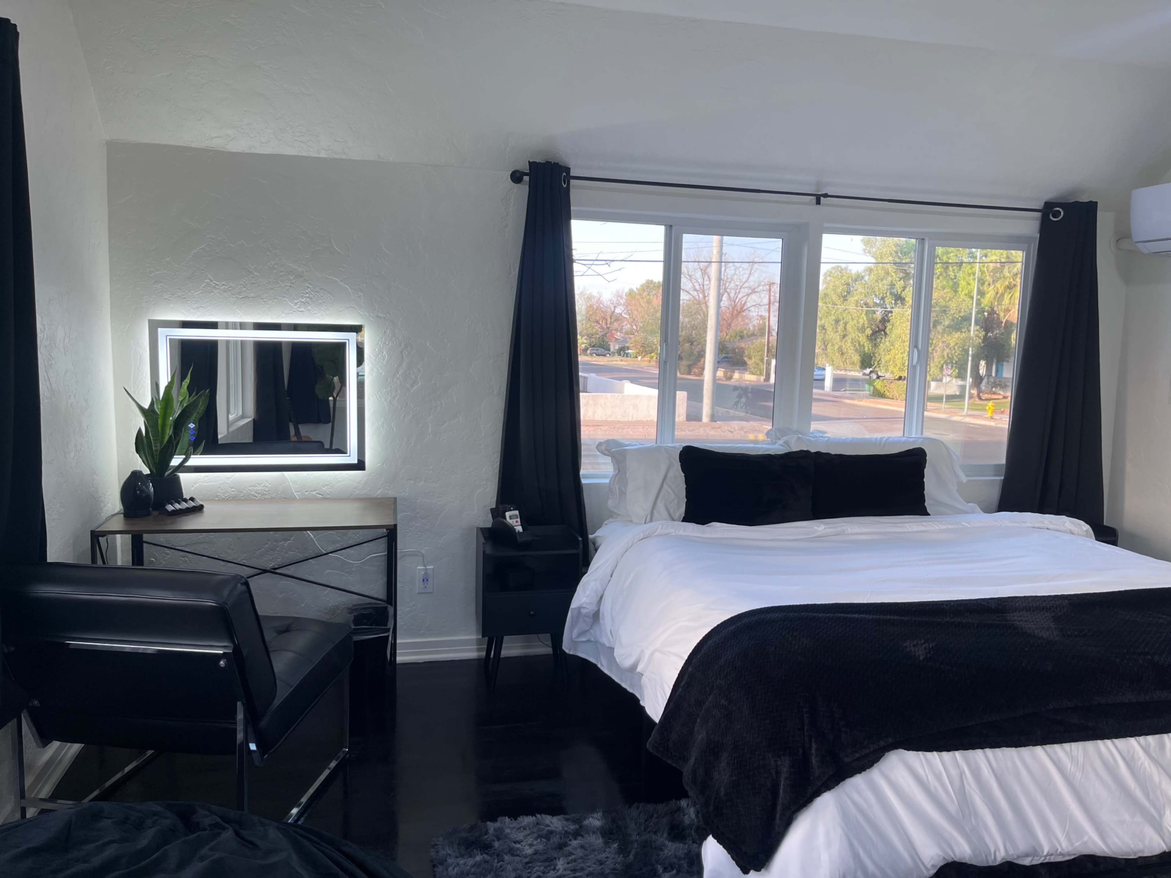 The image shows a modern bedroom featuring a bed with a white comforter, a mirrored vanity with a chair, and large windows allowing natural light to enter the space.