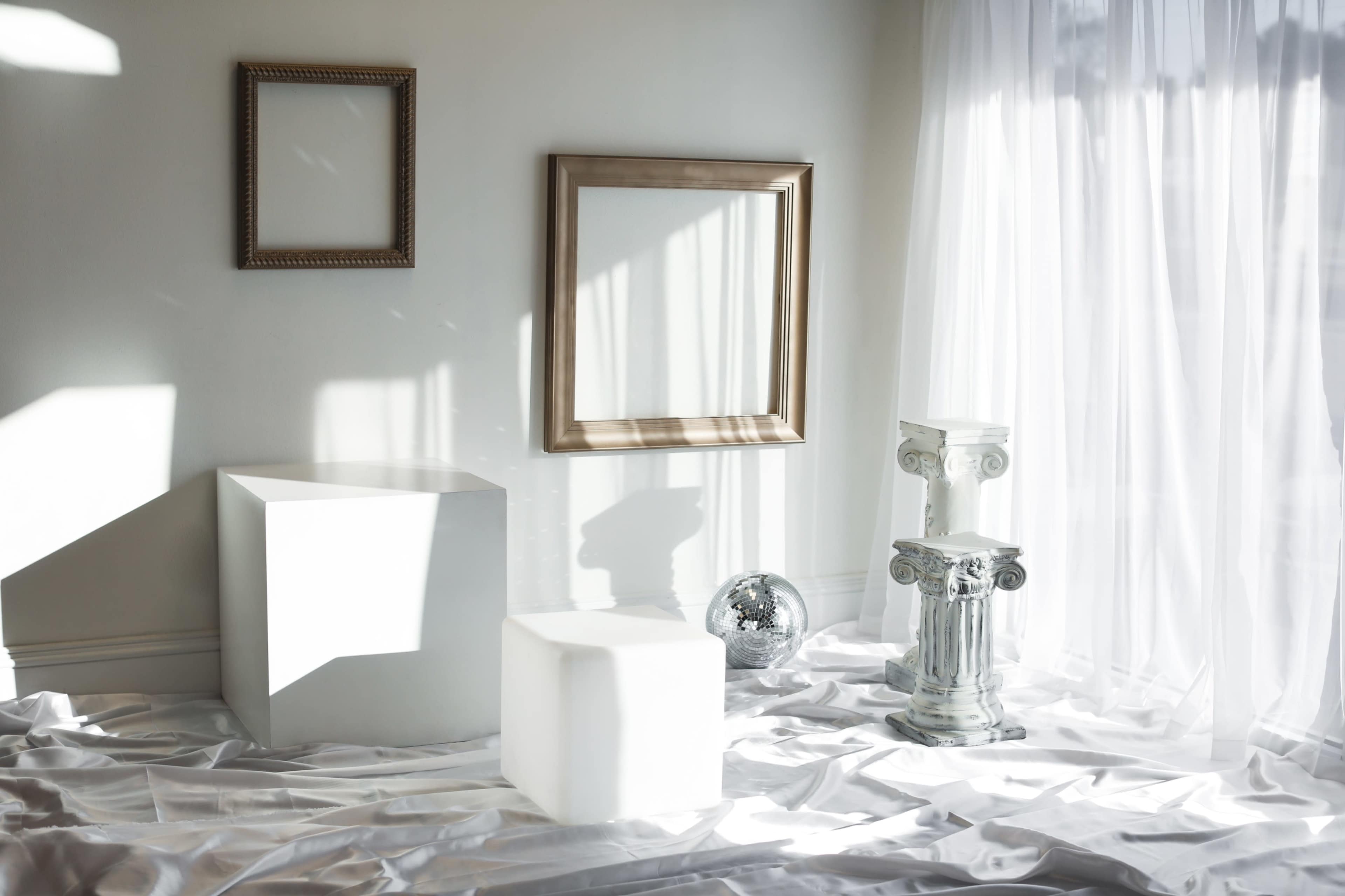 The scene features minimalist staging with two geometric white blocks, a classical column, a reflective sphere, and an ornate picture frame against a bright, airy backdrop.