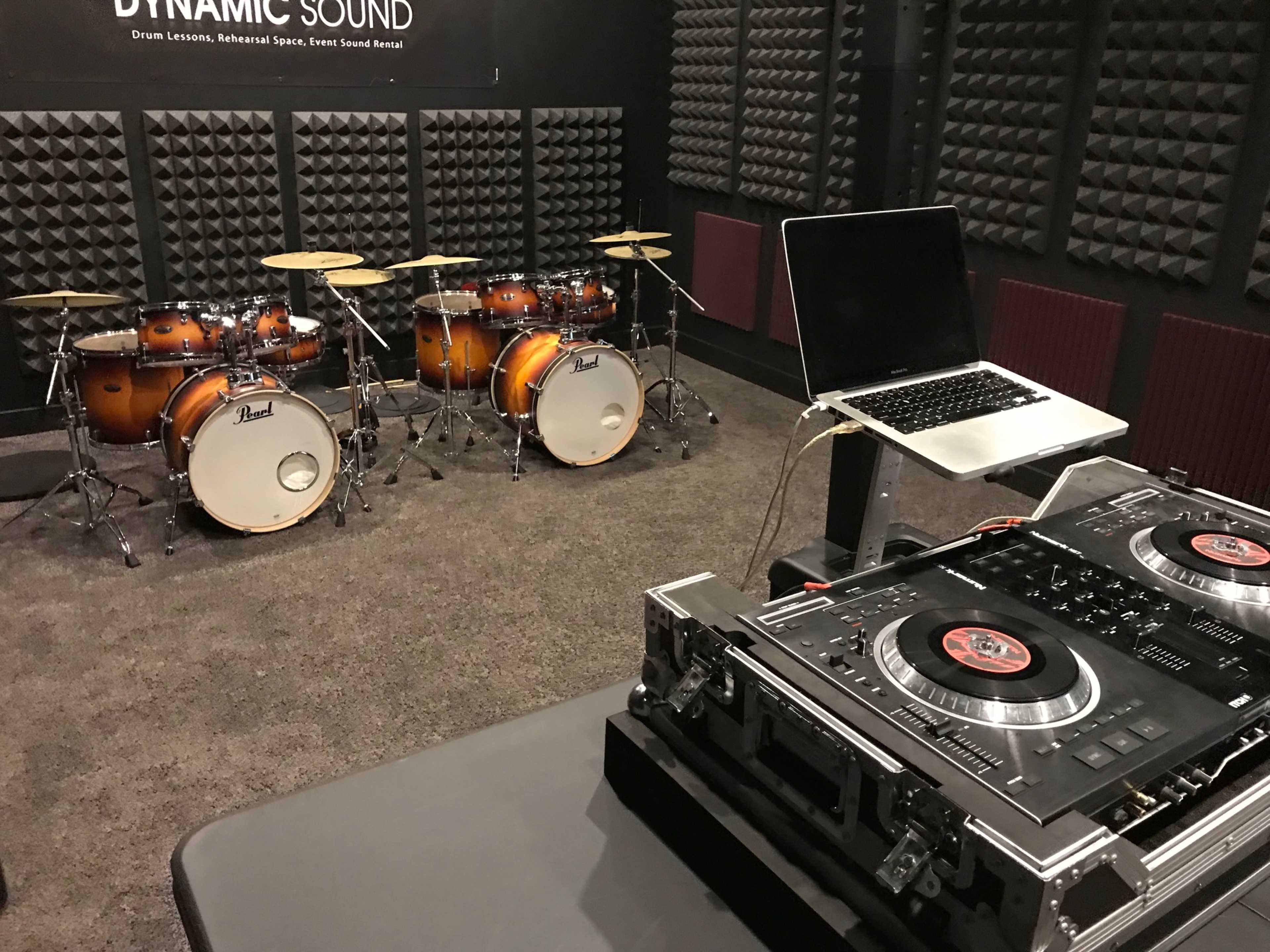 A music studio equipped with two drum sets, a laptop on a table, and a DJ turntable setup.