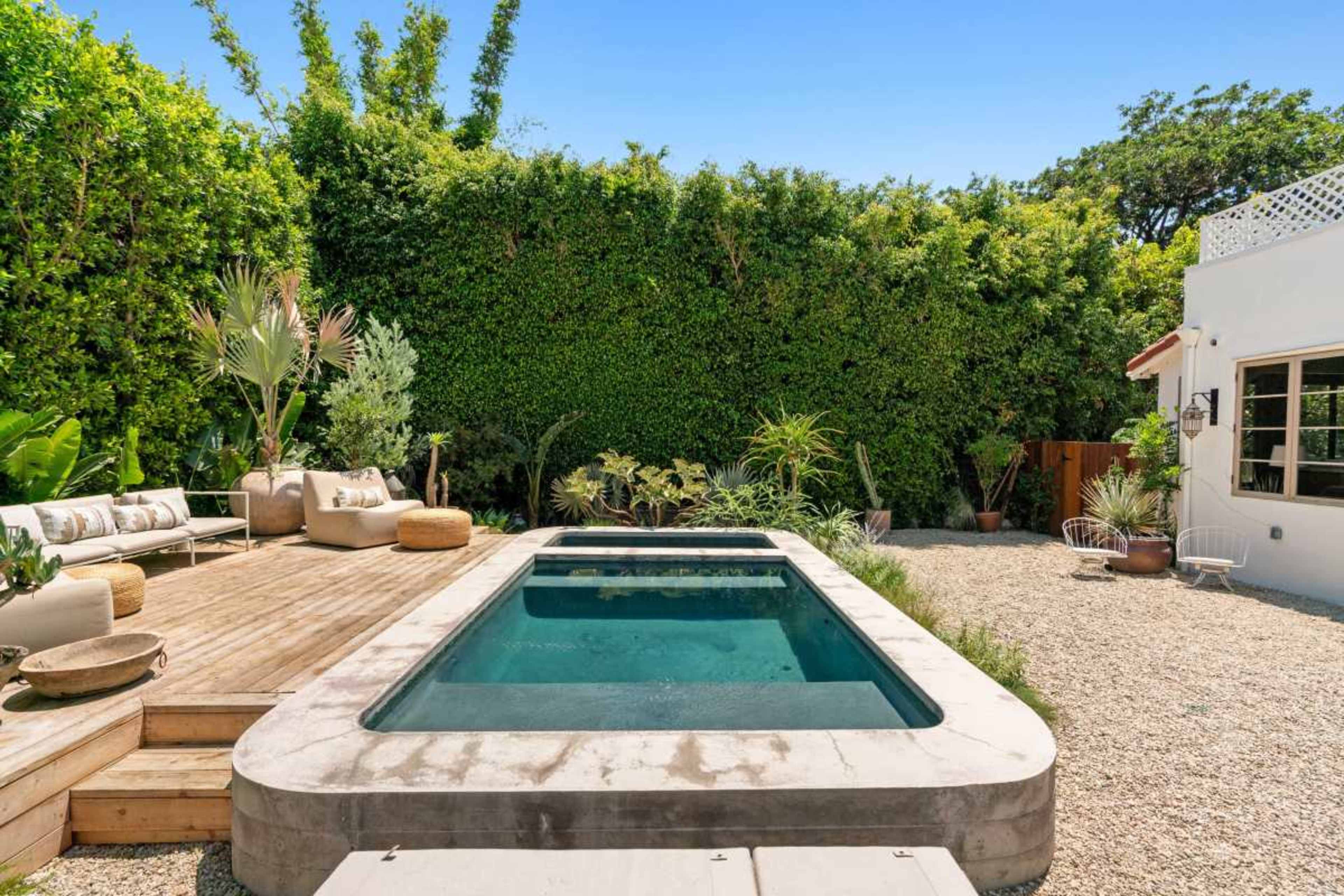 The image shows a backyard with a rectangular swimming pool surrounded by wooden decking, gravel, and tropical plants.