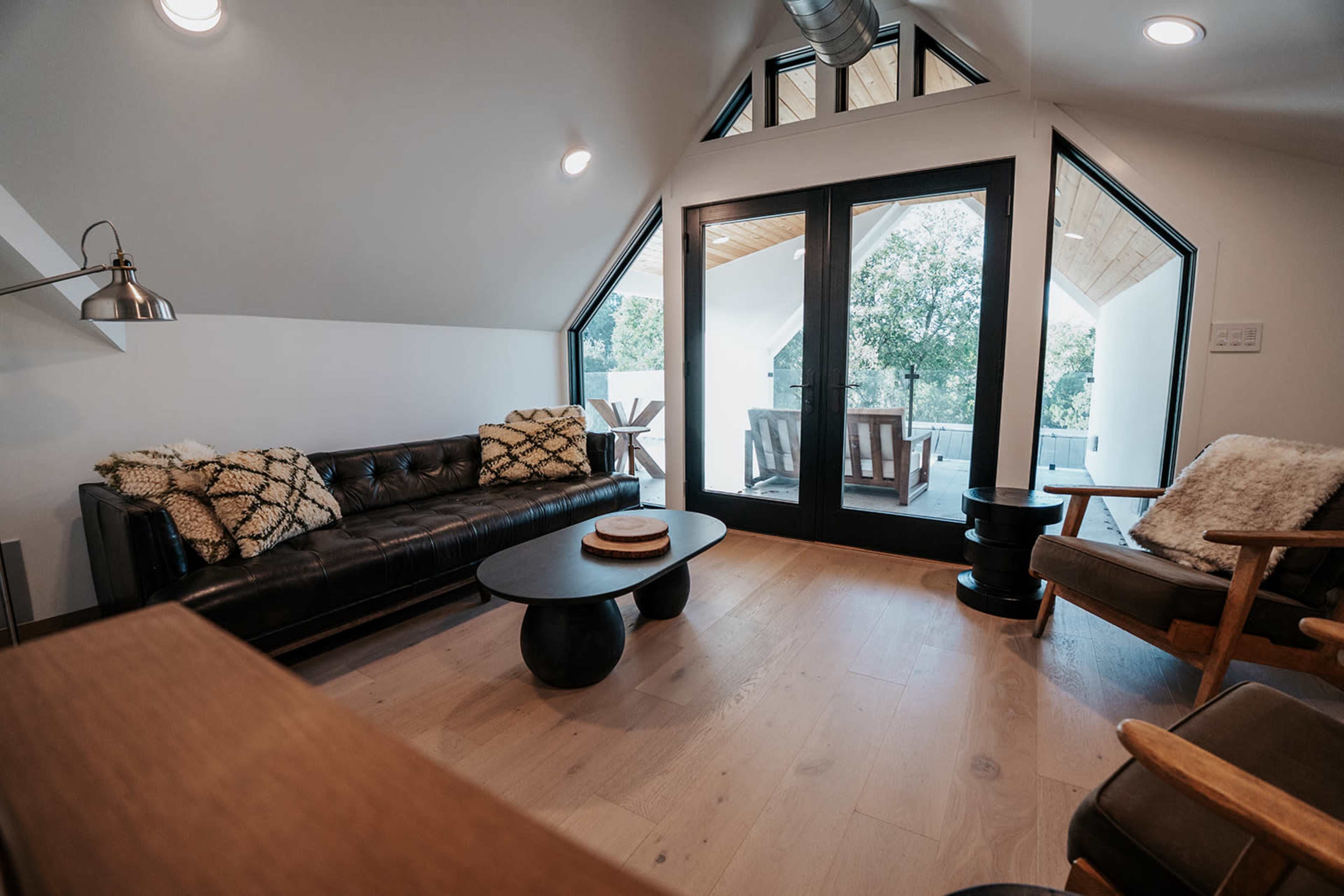 The image shows a modern living room with a black leather couch, a round coffee table, and large windows that let in natural light.