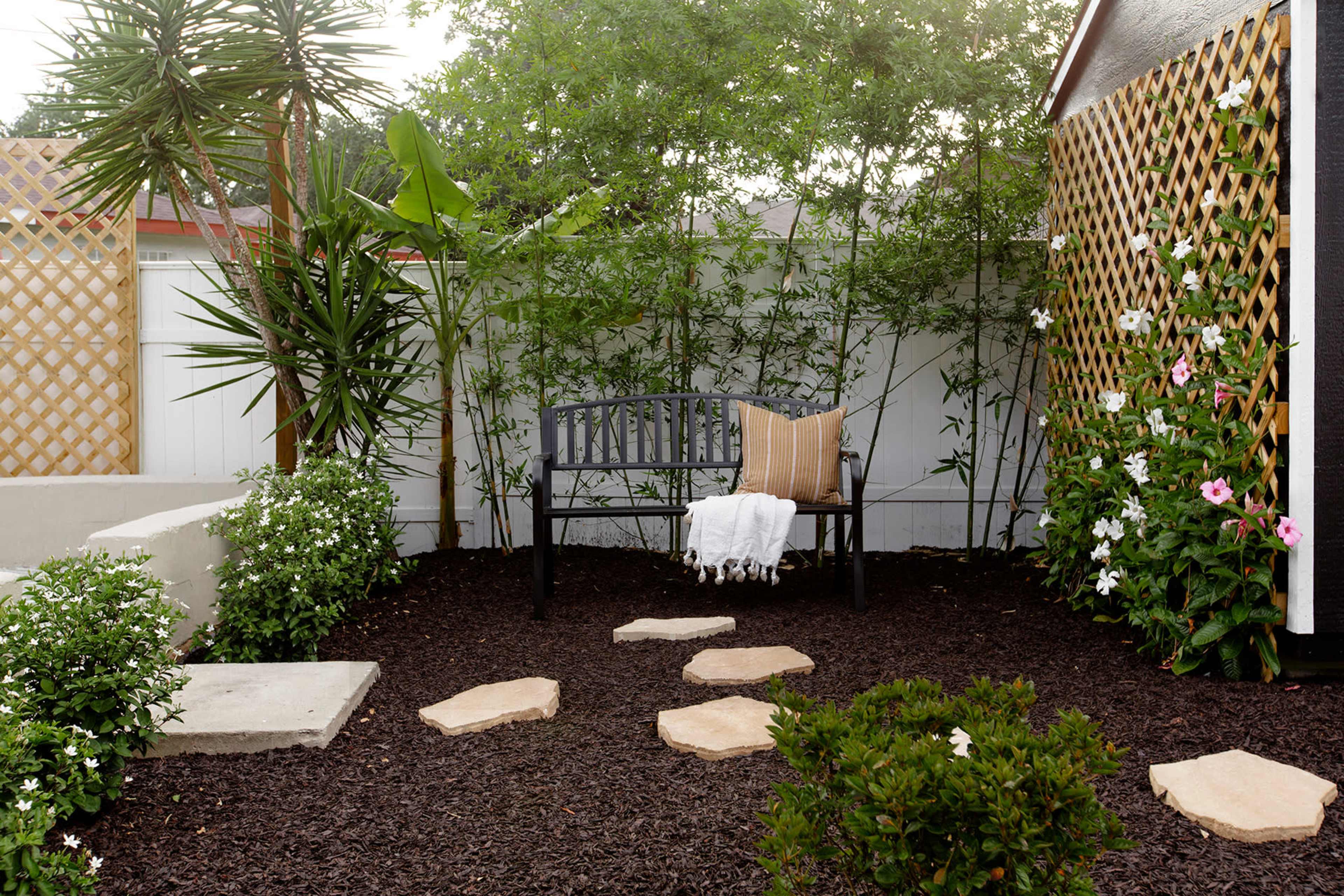 A wooden bench is placed on dark mulch in a small garden, with stone stepping paths and various plants surrounding it.