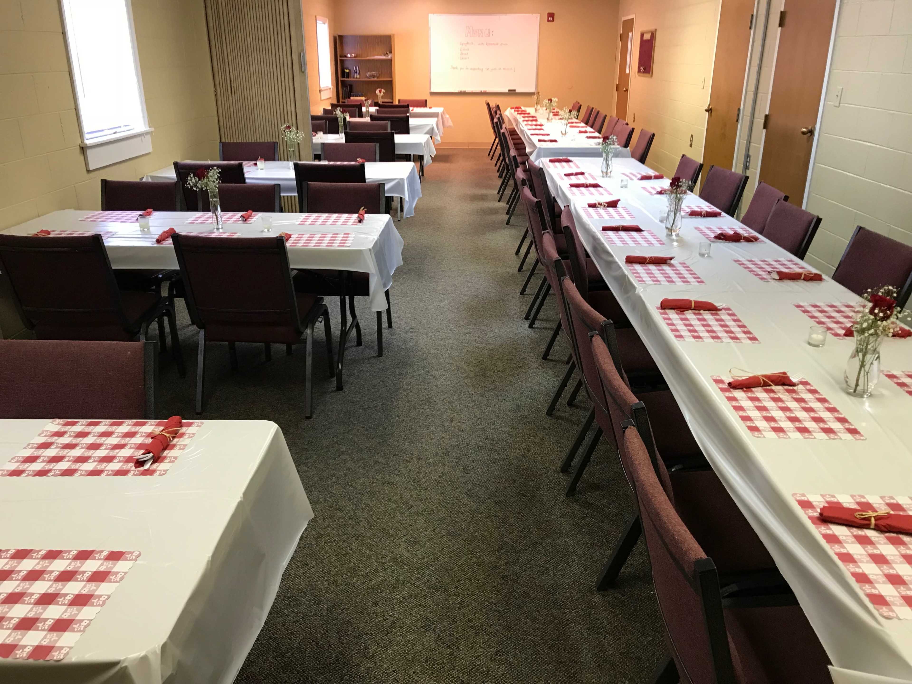 The image shows a neat dining setup with rows of tables covered in white cloth and red-checkered placemats, arranged in a spacious room.